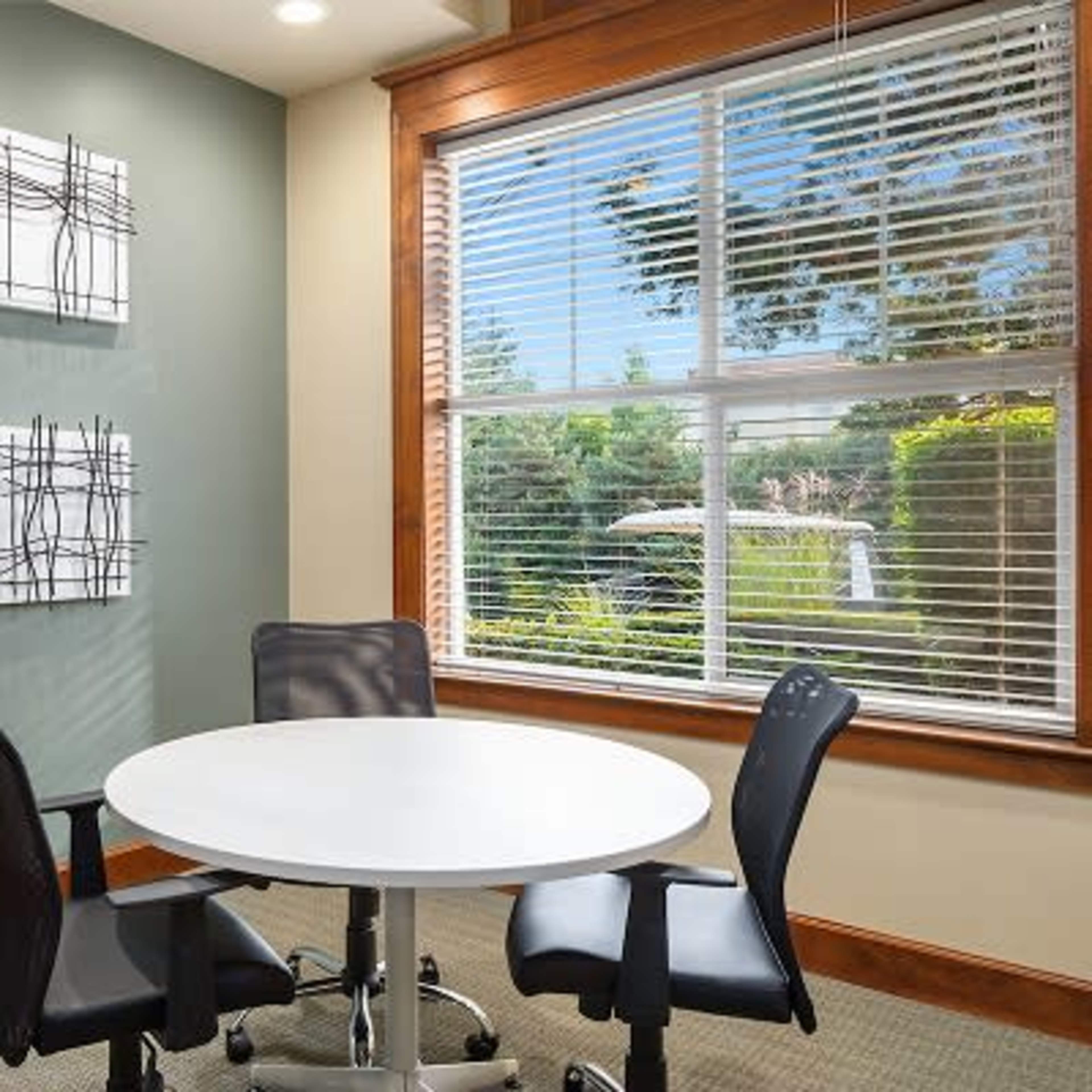 A round white table surrounded by two black chairs is positioned beside a window with blinds, offering a view of greenery outside, while three abstract wall art pieces are displayed on the green wall.