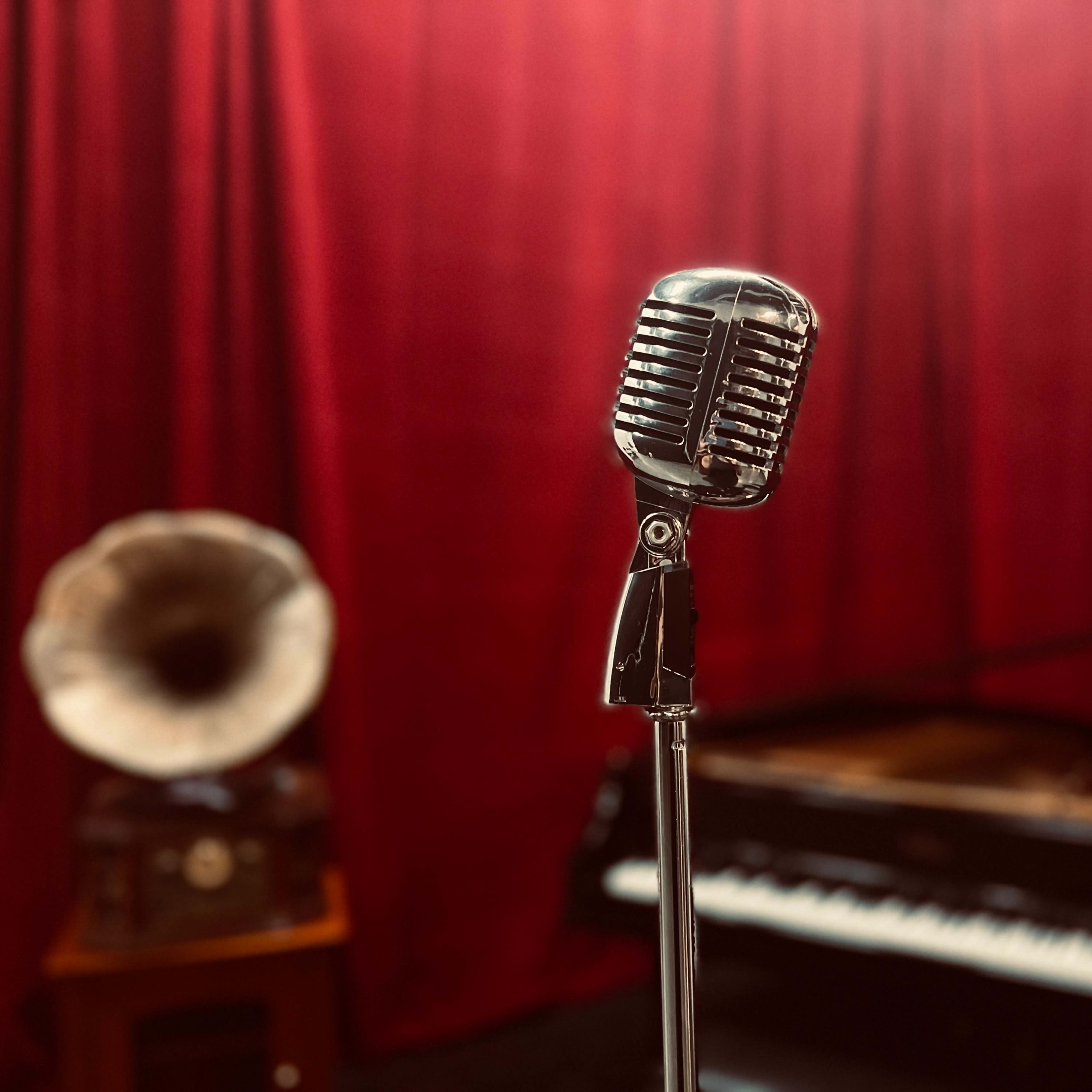 A vintage microphone stands in front of a red curtain, with a grand piano and a gramophone visible in the background.