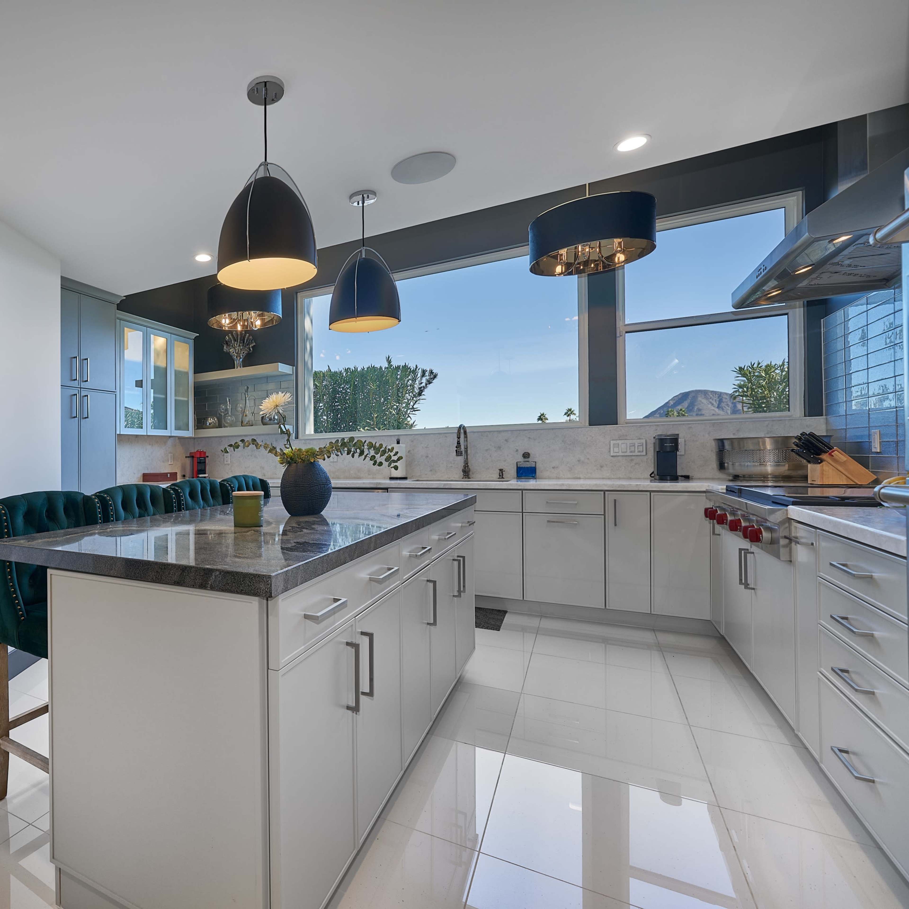 A modern kitchen features white cabinetry, a large island with a gray countertop, and pendant lights hanging above, with large windows providing natural light and views.
