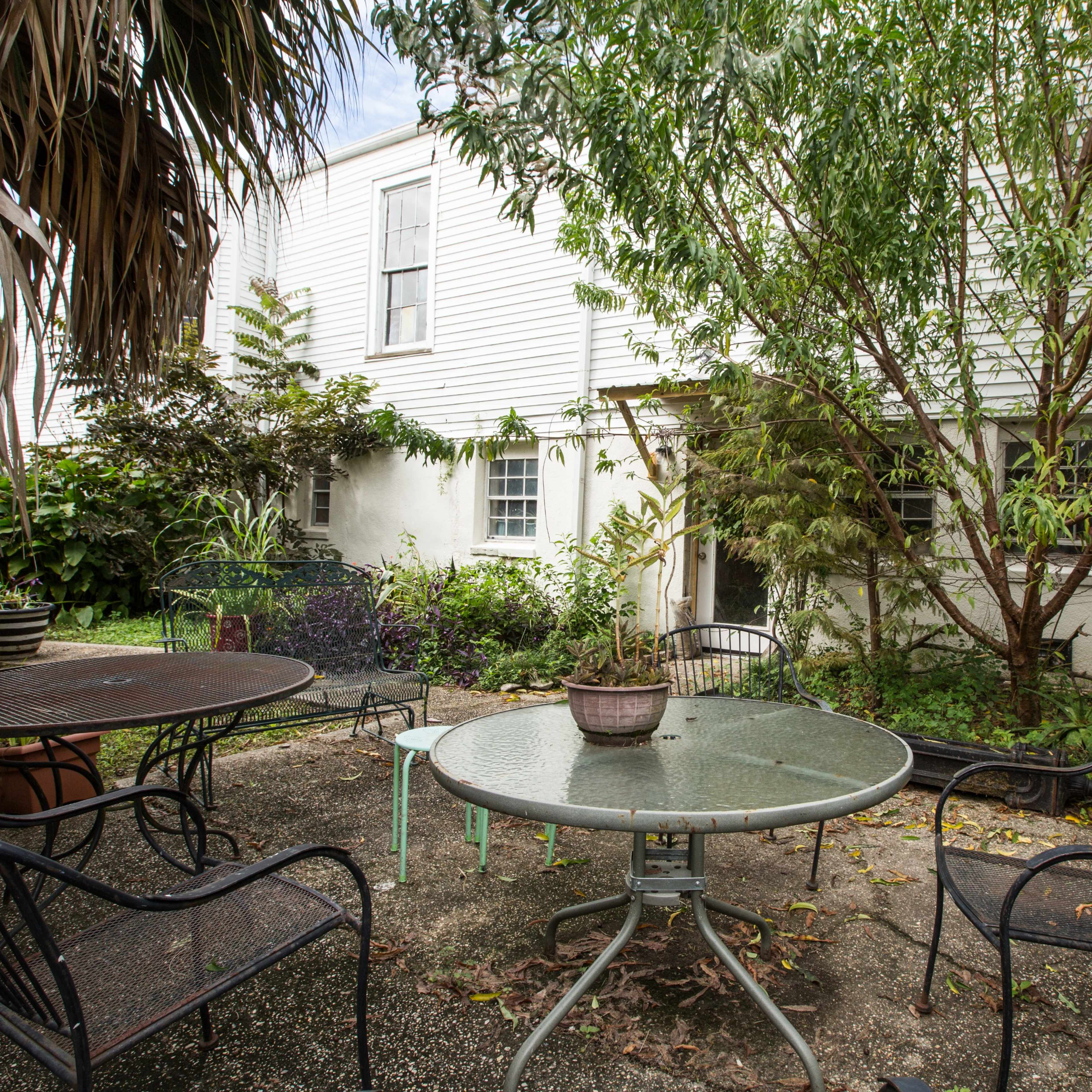 A small outdoor space with two metal tables and several chairs surrounded by plants and a white building in the background.