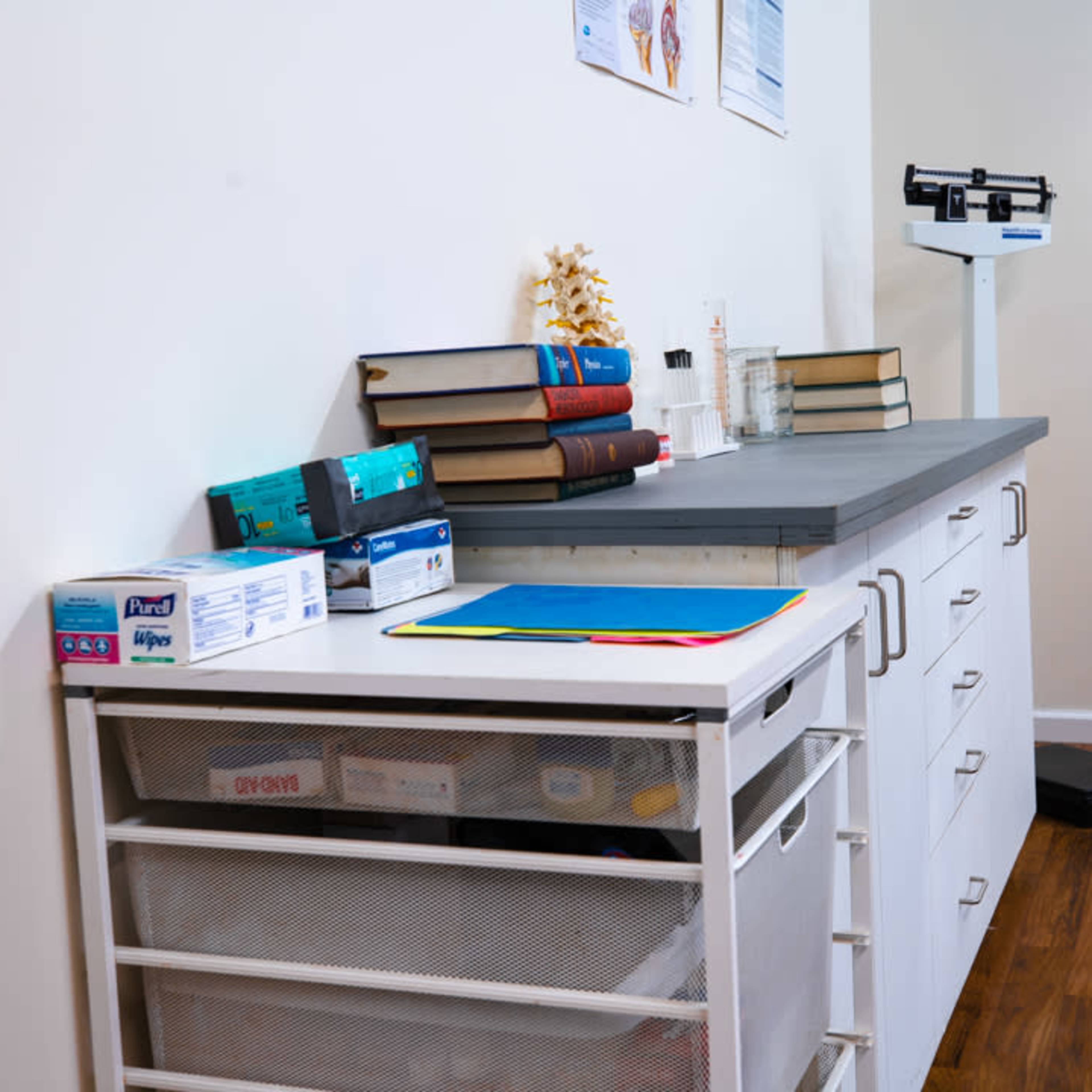A medical examination room features a countertop with stacked books, a spine model, and medical supplies, alongside a scale and storage drawers.