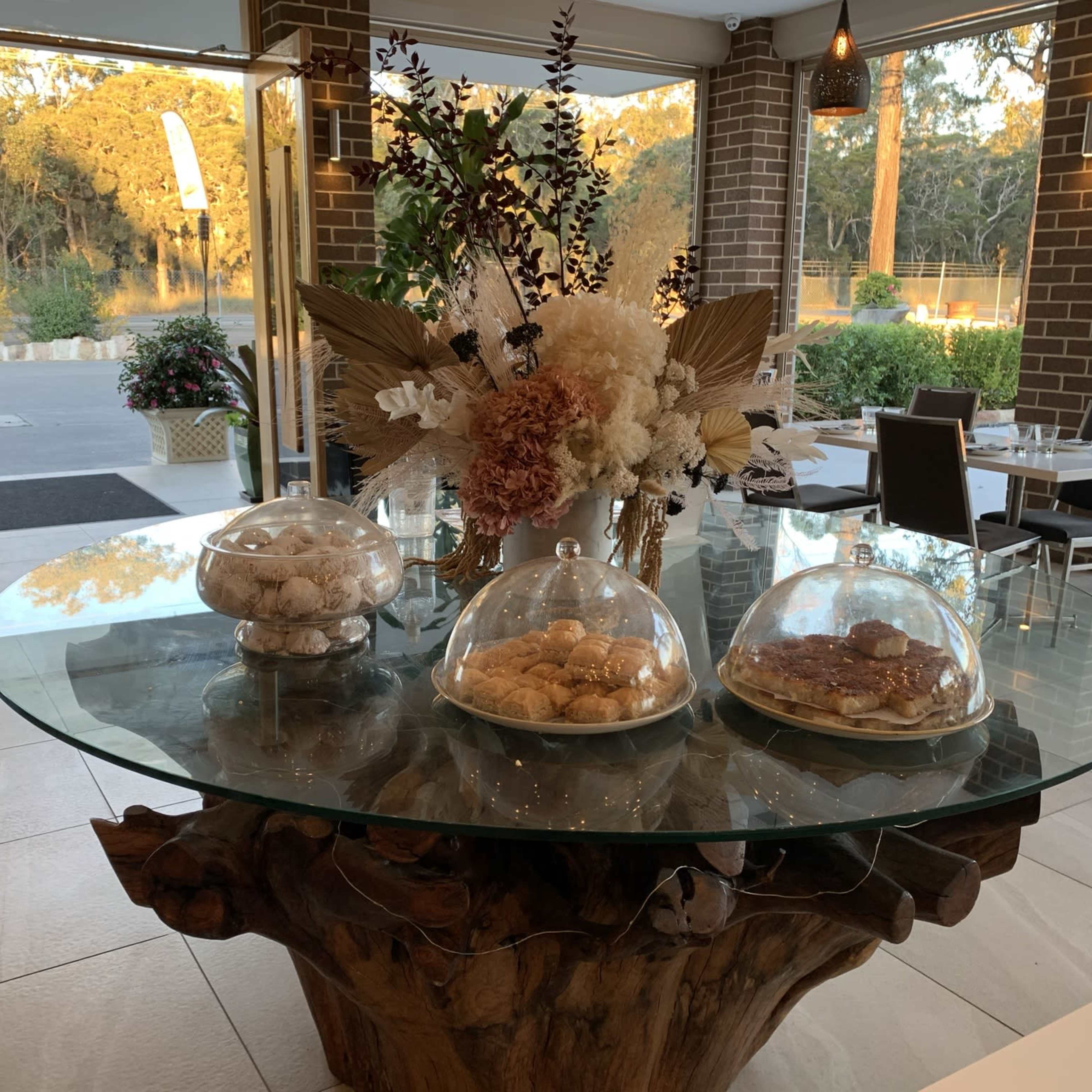 A glass table featuring assorted pastries displayed under cloches is set in a warmly lit restaurant with large windows overlooking greenery.