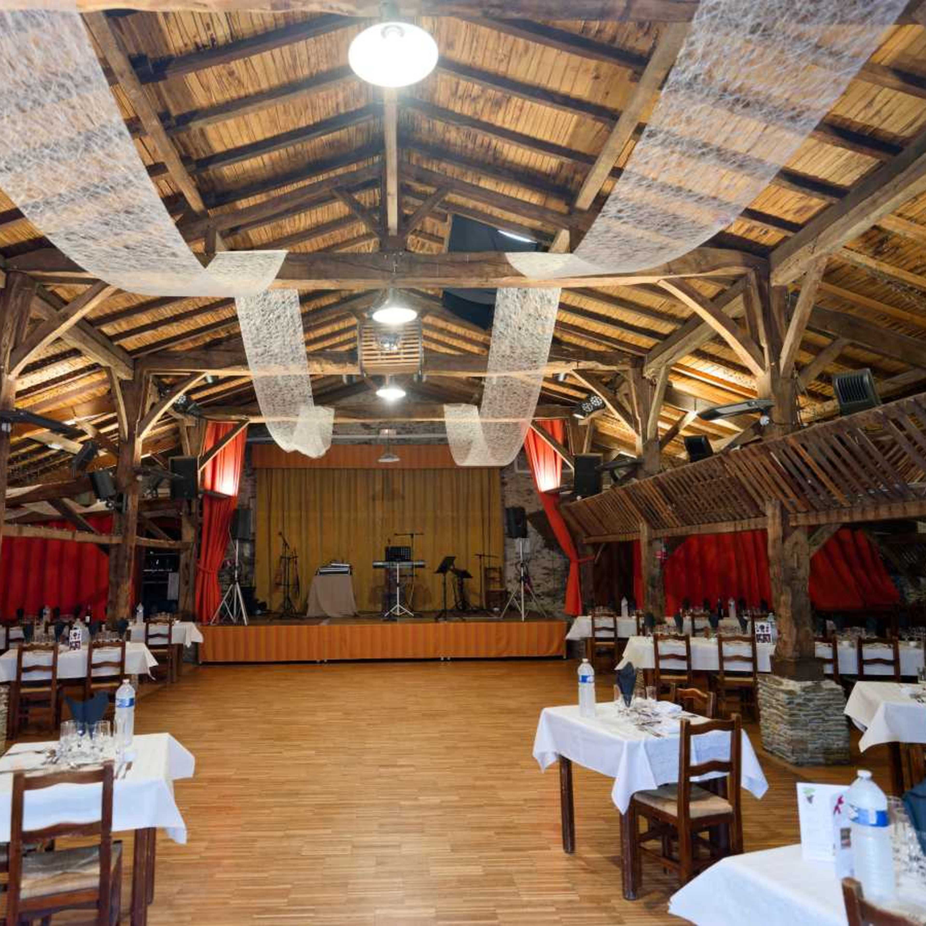 A rustic restaurant interior with wooden beams, tables set with white tablecloths, and a small stage at the front.