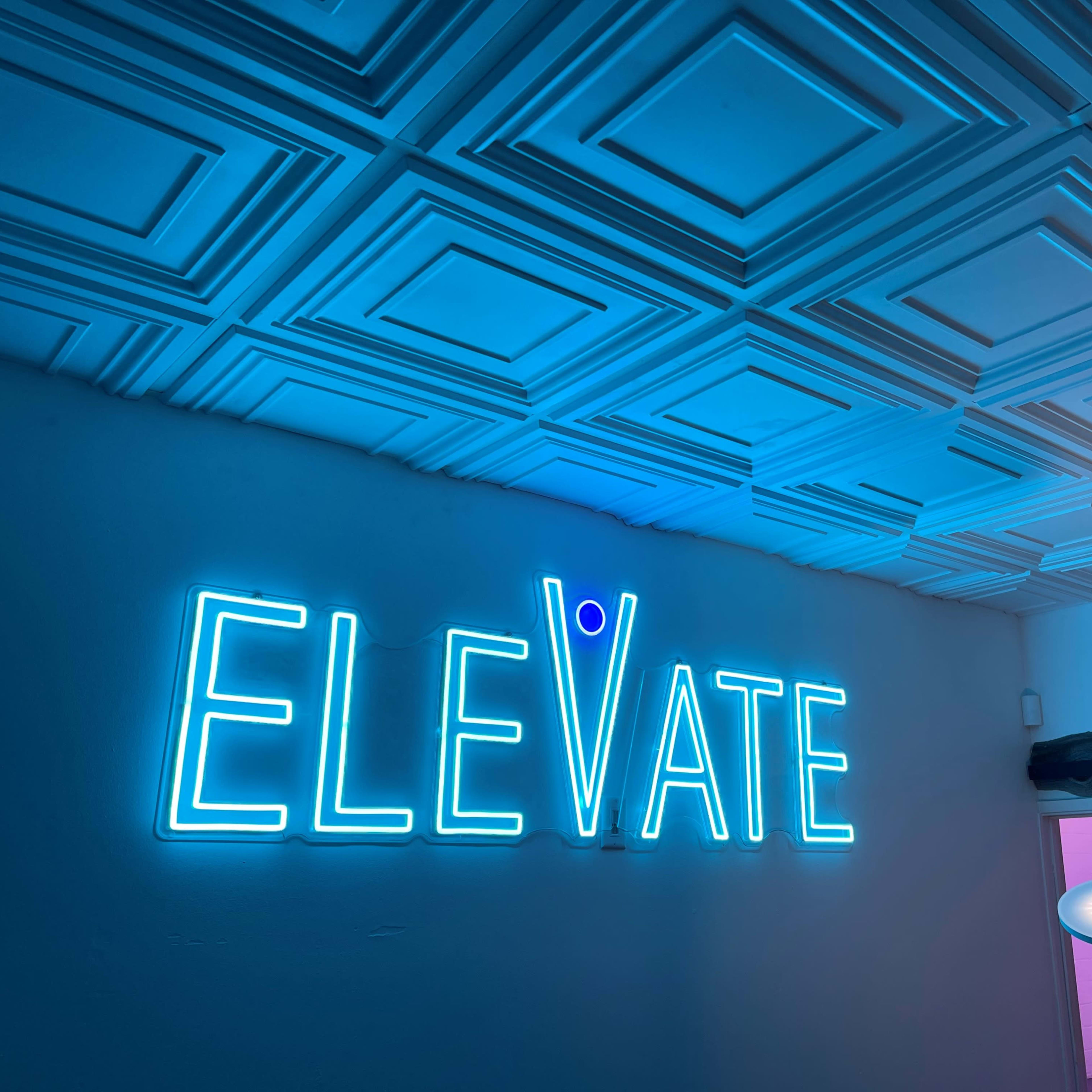 A bright blue neon sign displaying the word "ELEVATE" is mounted on a white wall with a decorative ceiling above.