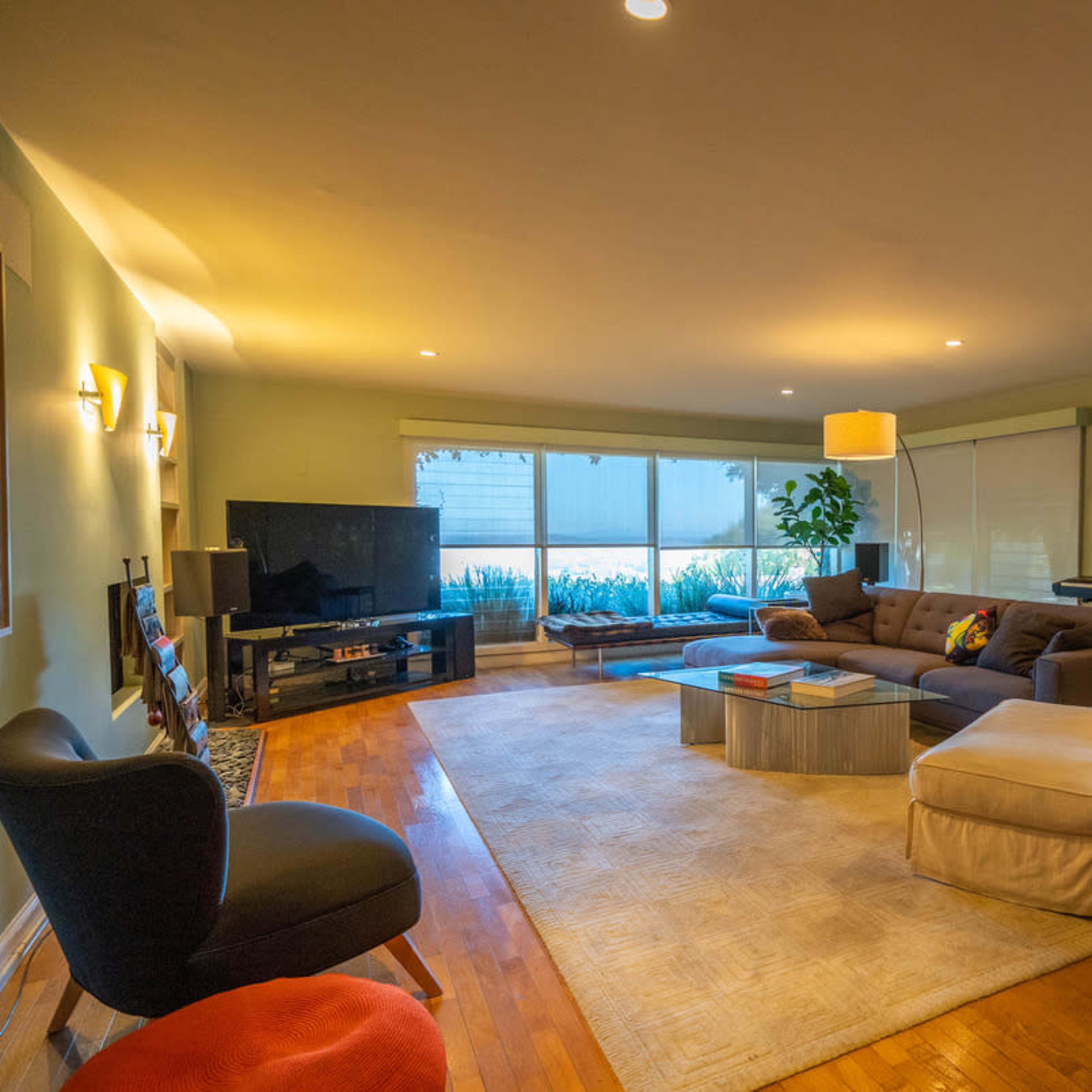 The room features a spacious living area with a sectional sofa, a large television, and windows allowing natural light, complemented by a green wall and artwork.