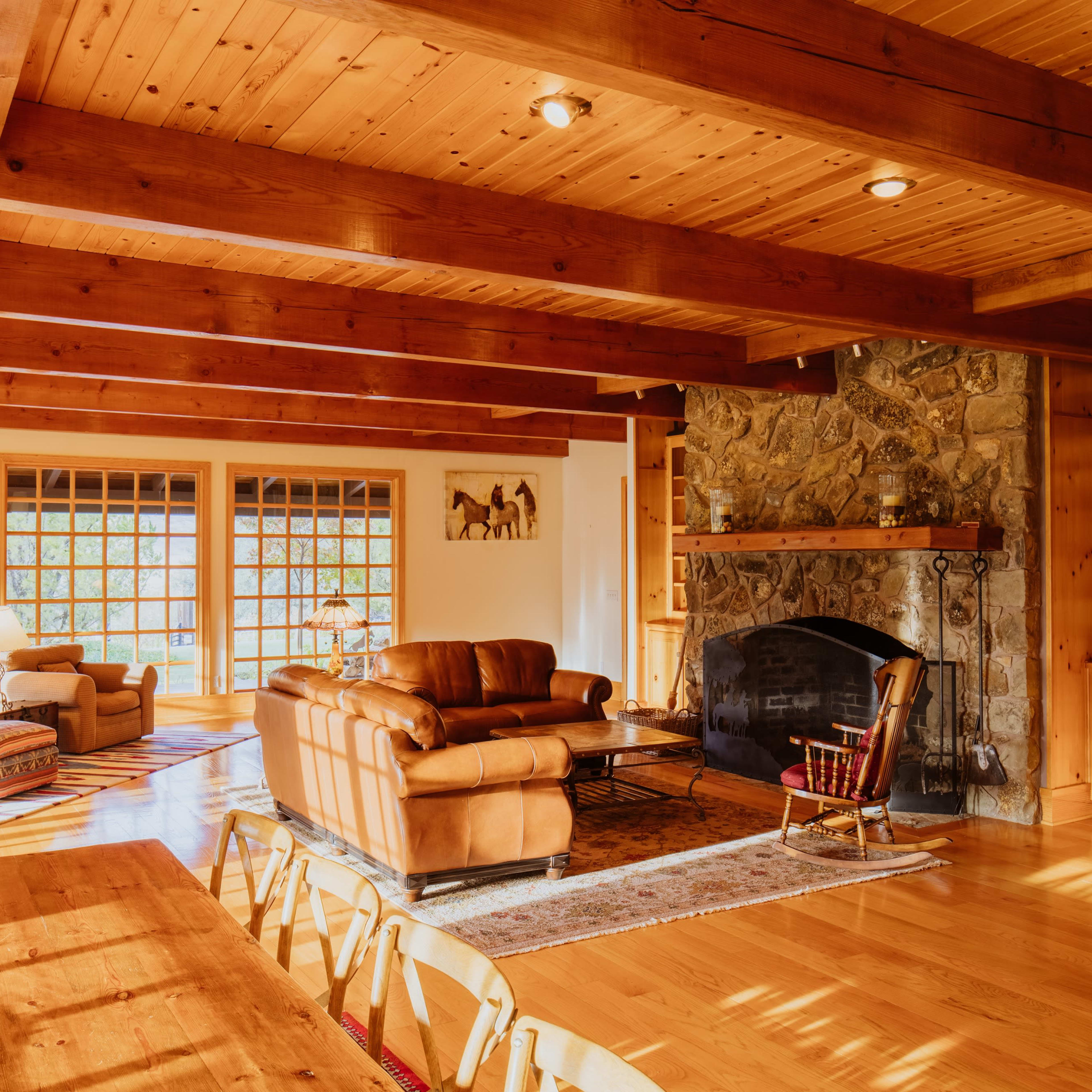 A cozy living room with wooden beams, a stone fireplace, and large windows that allow natural light to fill the space.