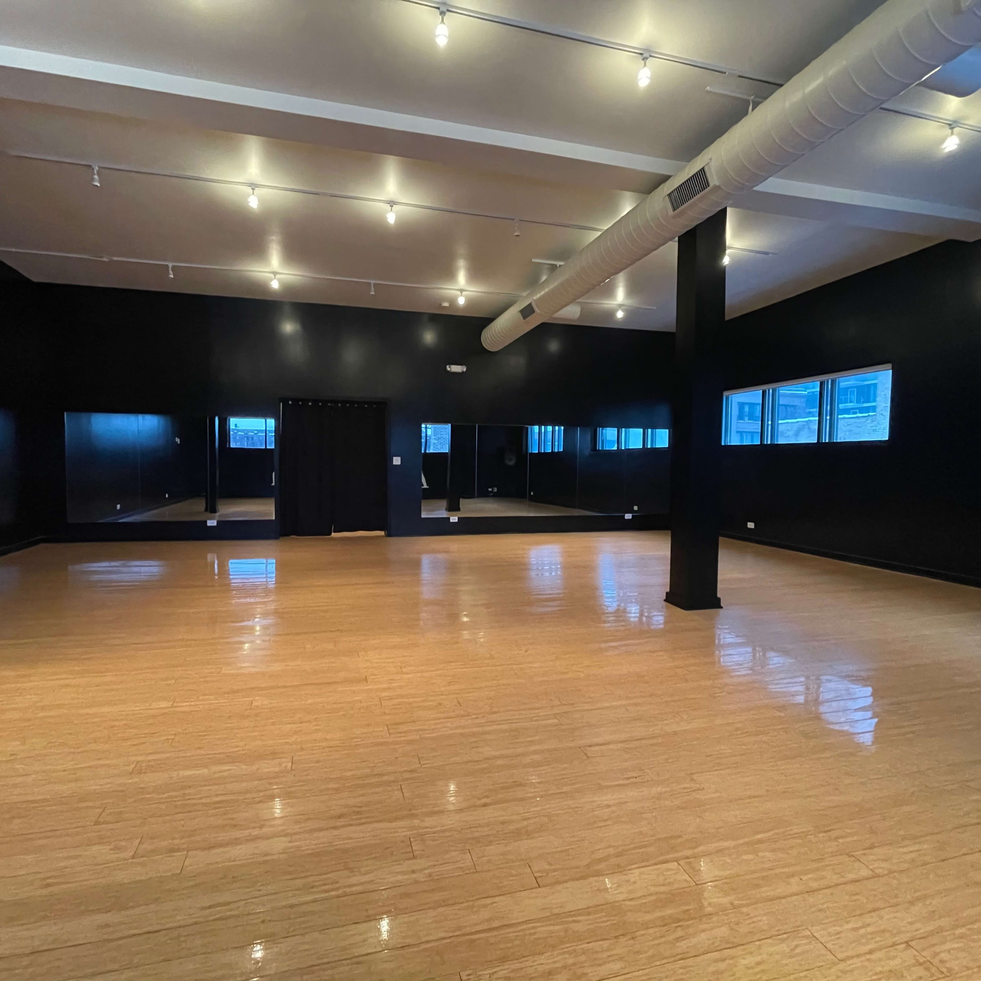 A spacious dance studio with a wooden floor, black walls, and large windows allowing natural light.