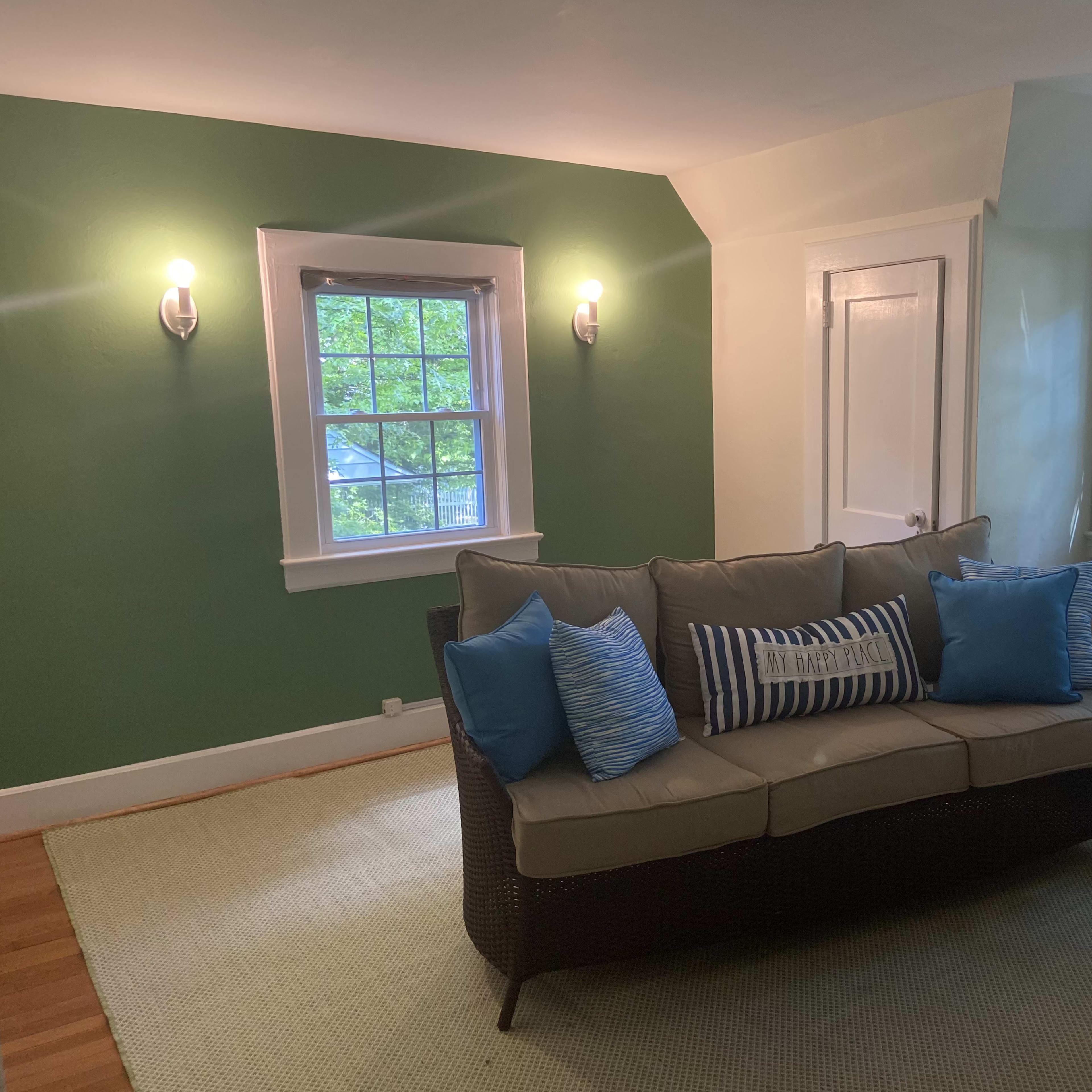 The scene shows a living room with a green accent wall, a light-colored sofa adorned with blue decorative pillows, and two windows allowing natural light to enter.