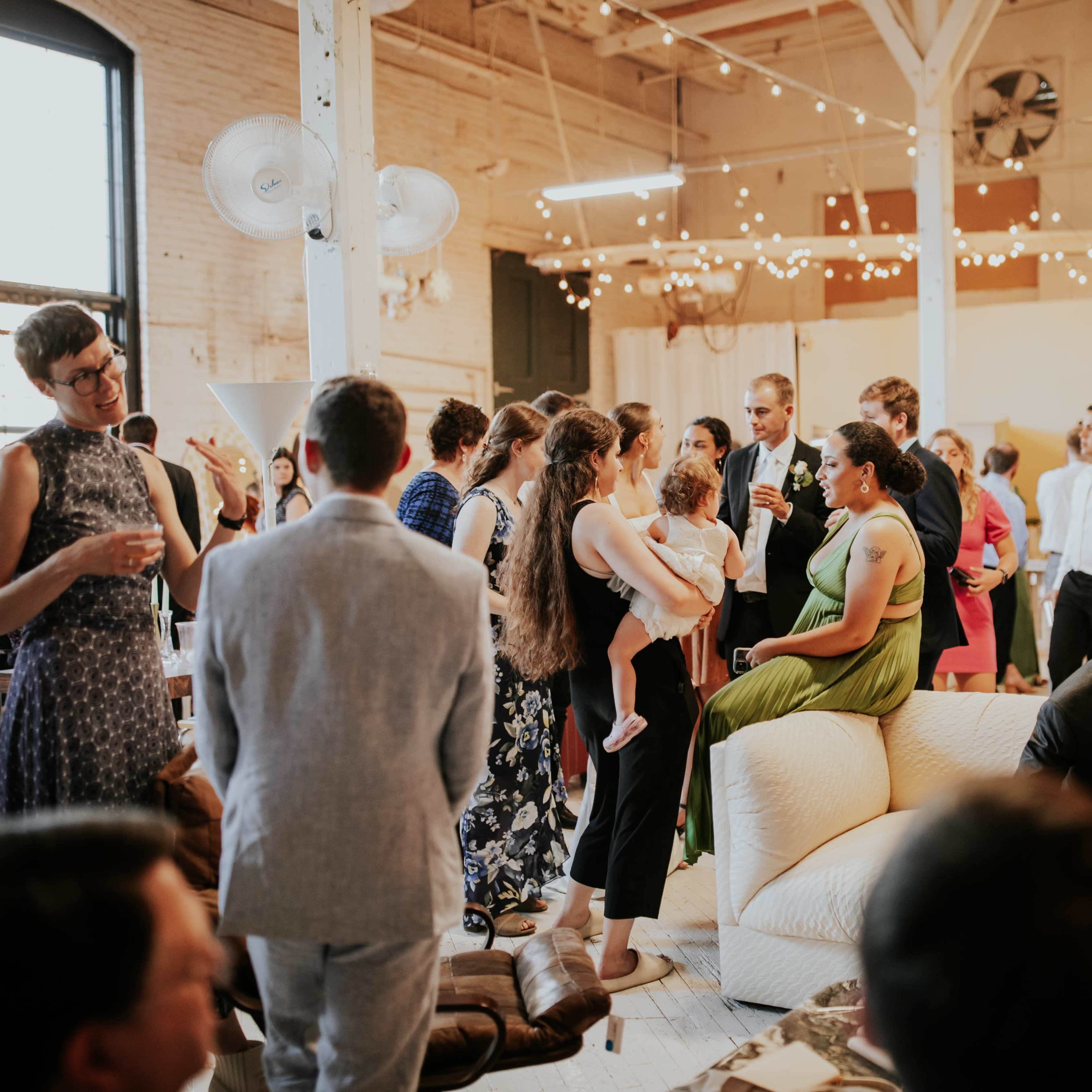 A large group of people socialize at a lively indoor event, surrounded by string lights and various seating arrangements.