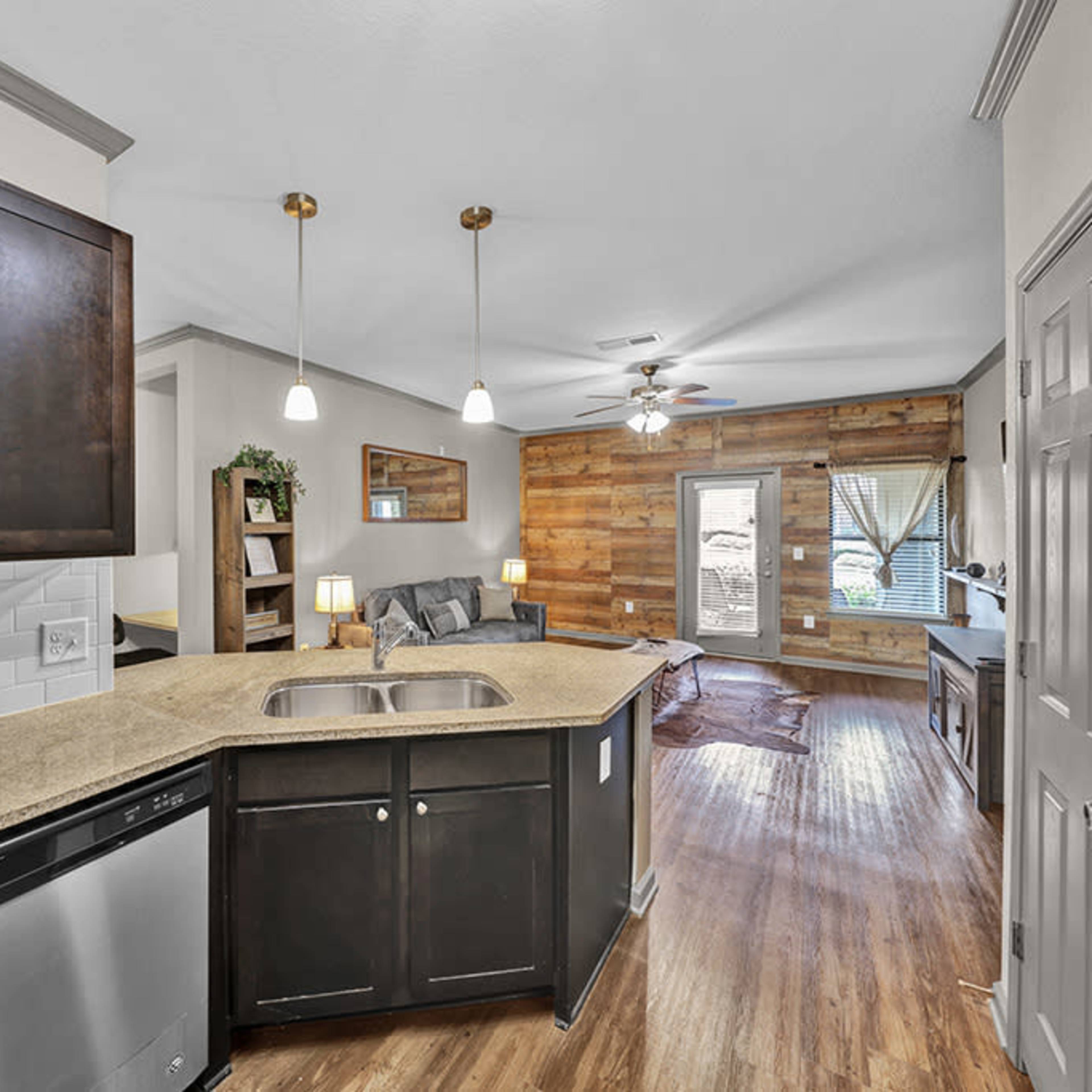 A kitchen with dark cabinets and a granite countertop, adjacent to a living area featuring a wooden accent wall and a door leading outside.