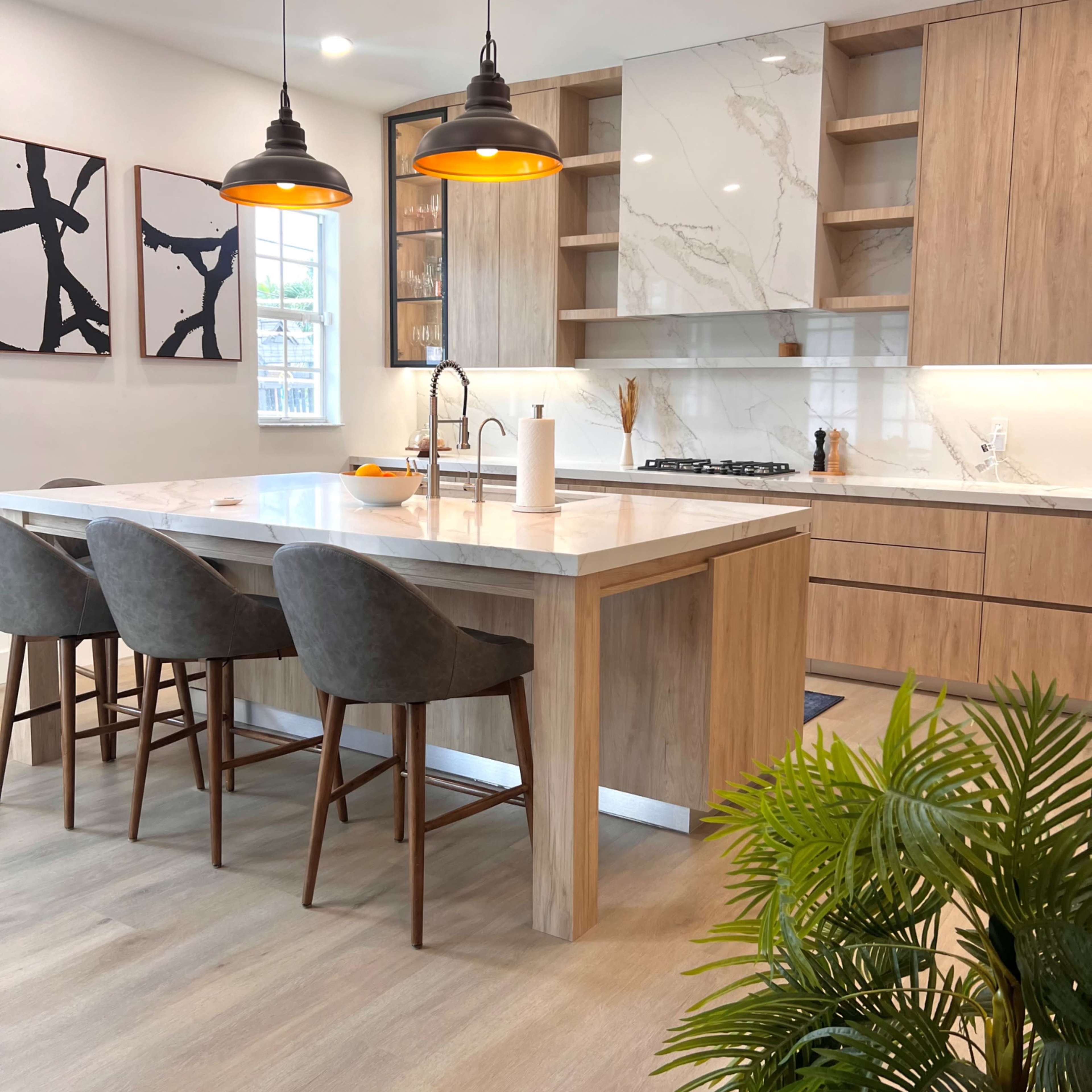 A modern kitchen features a large wooden island with four gray upholstered chairs, pendant lights overhead, and minimalist cabinetry alongside a marble backsplash.