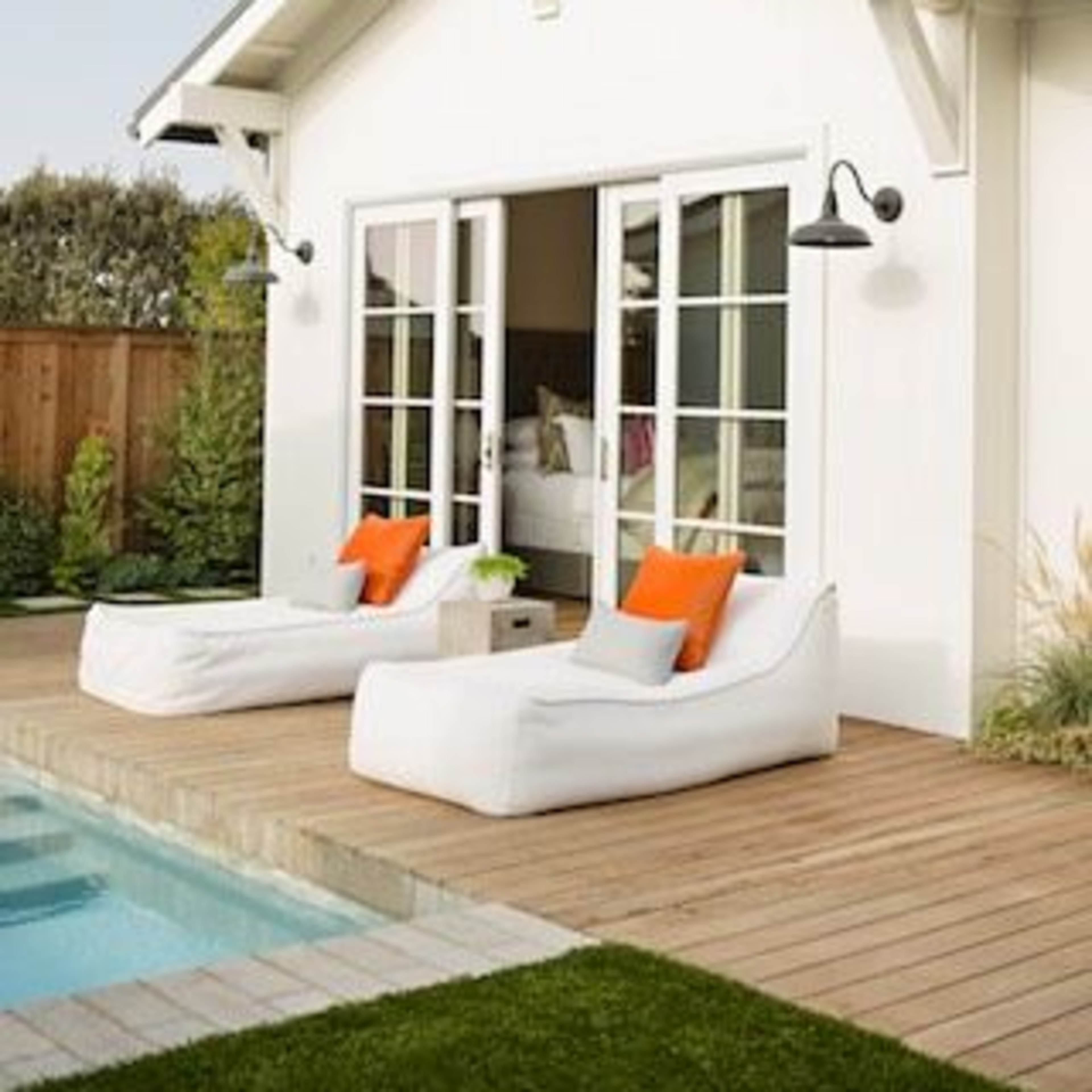 A modern white house features large sliding doors opening onto a wooden deck with two white lounge chairs and a swimming pool.
