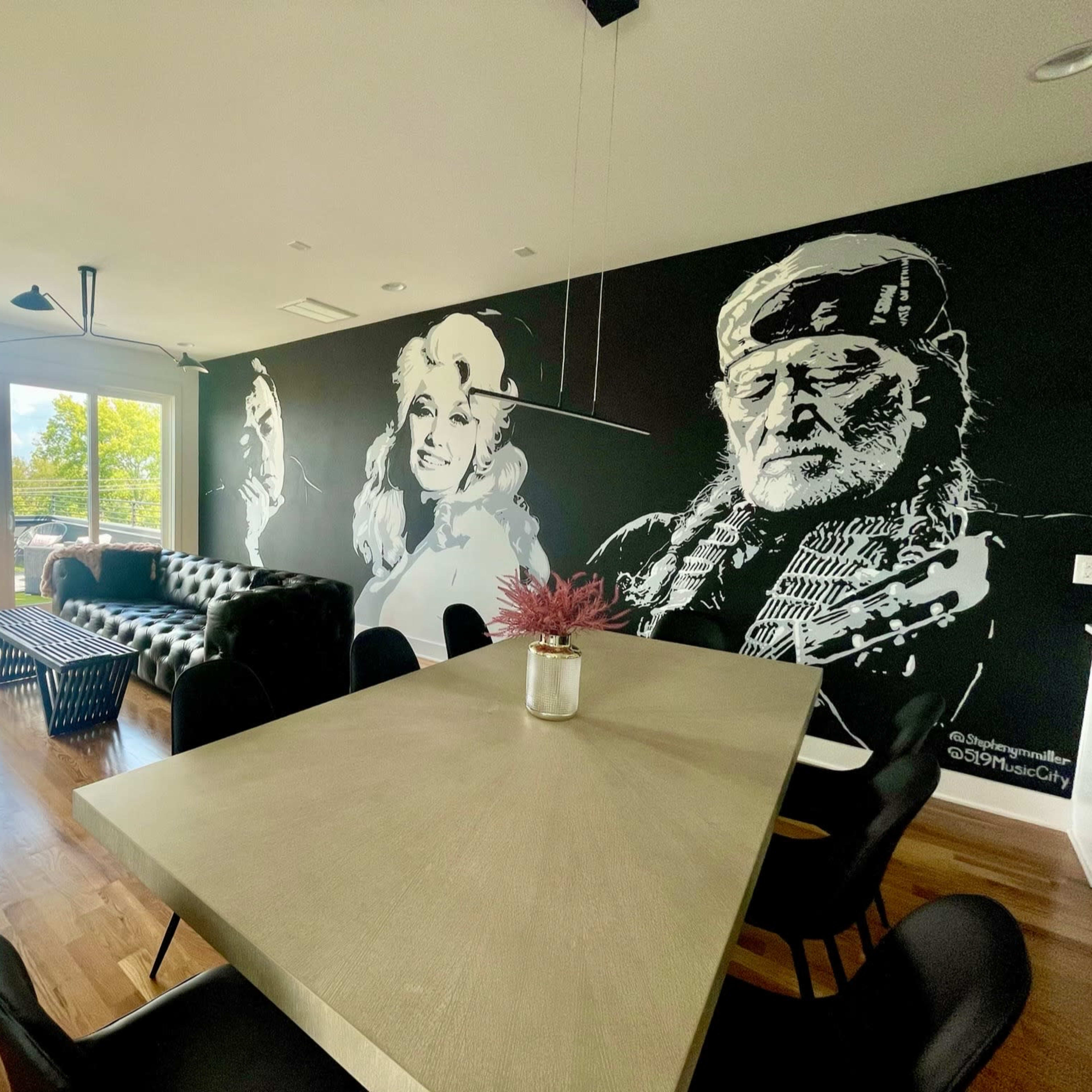 A modern living space featuring a mural of two figures on a black wall, with a dining table in the center and a view of the outdoors through large windows.