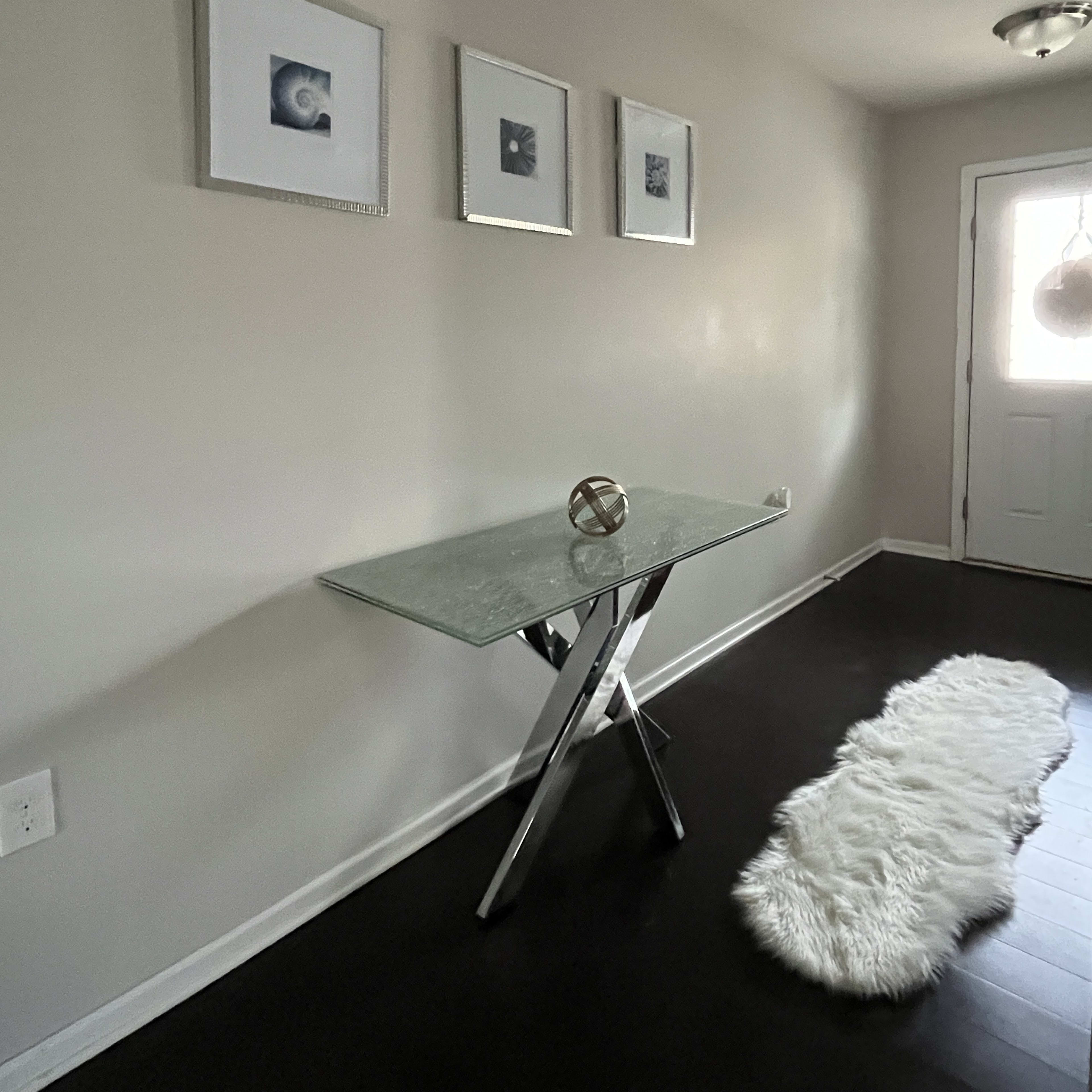 A glass-top table with a metallic base sits against a light-colored wall, accompanied by three framed pictures and a white faux-fur runner leading to a door.