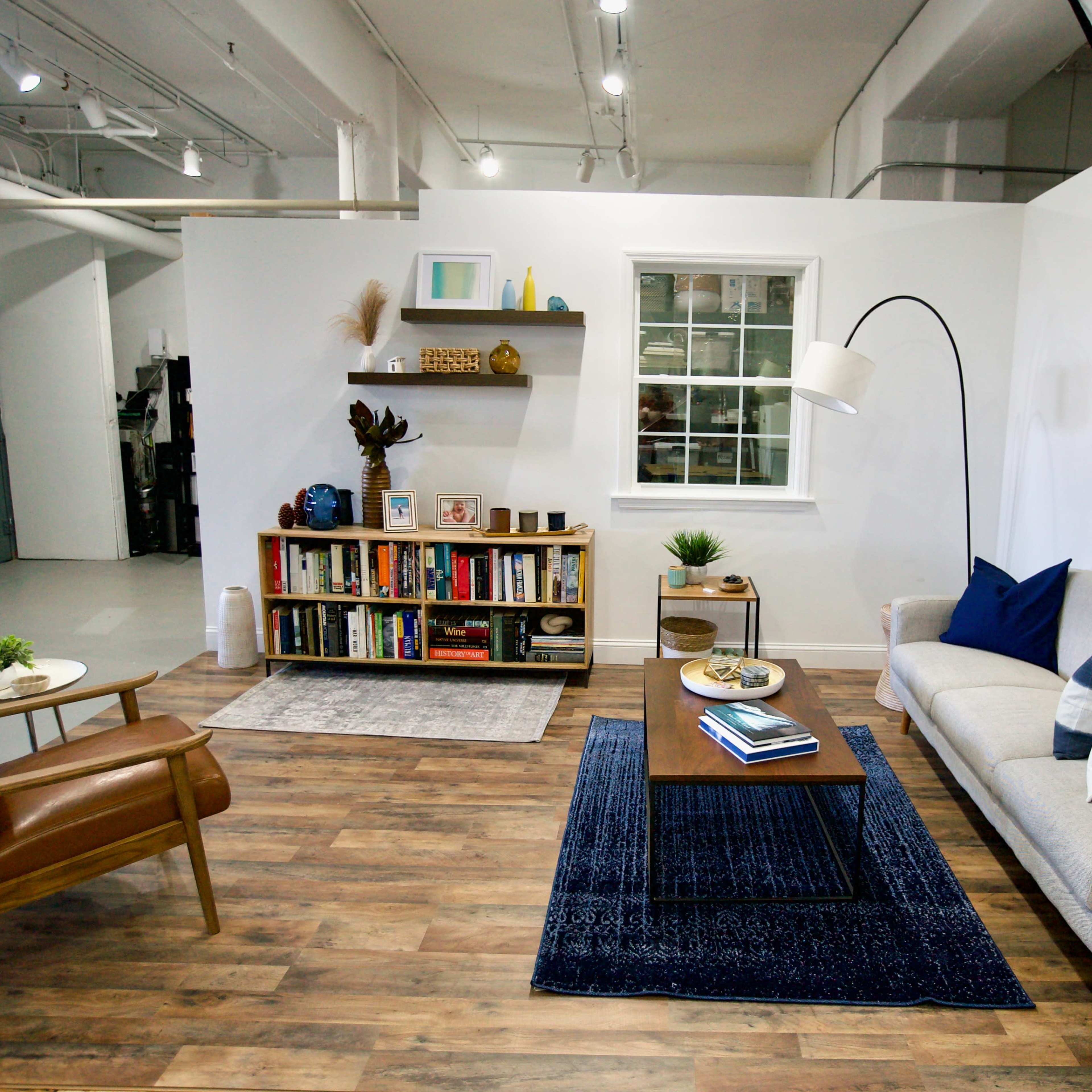 A modern living room setup with a beige sofa, a wooden coffee table on a blue rug, a bookshelf filled with books, and a mid-century modern chair.