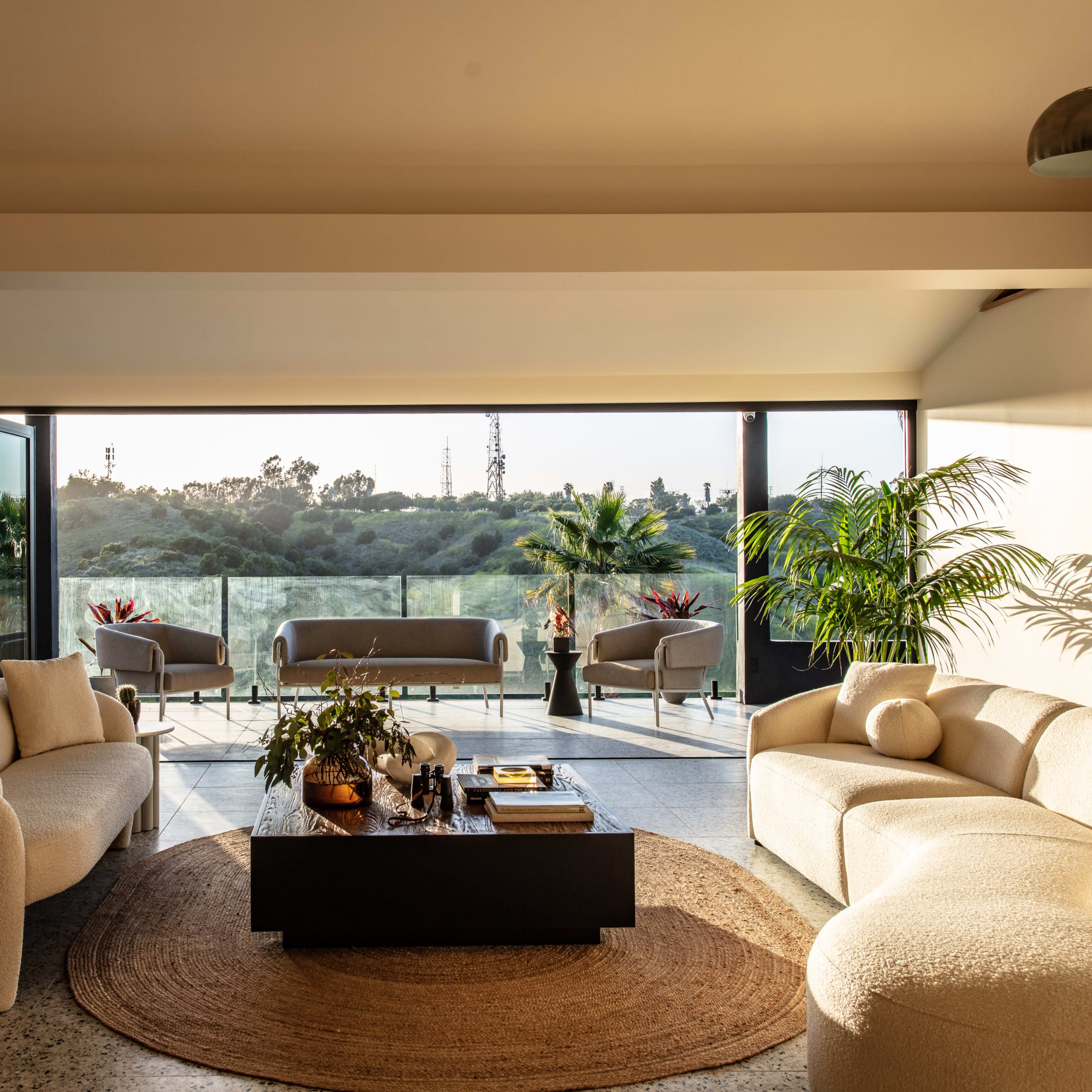 A modern living room features curved sofas and a coffee table overlooking a scenic balcony with greenery in the background.