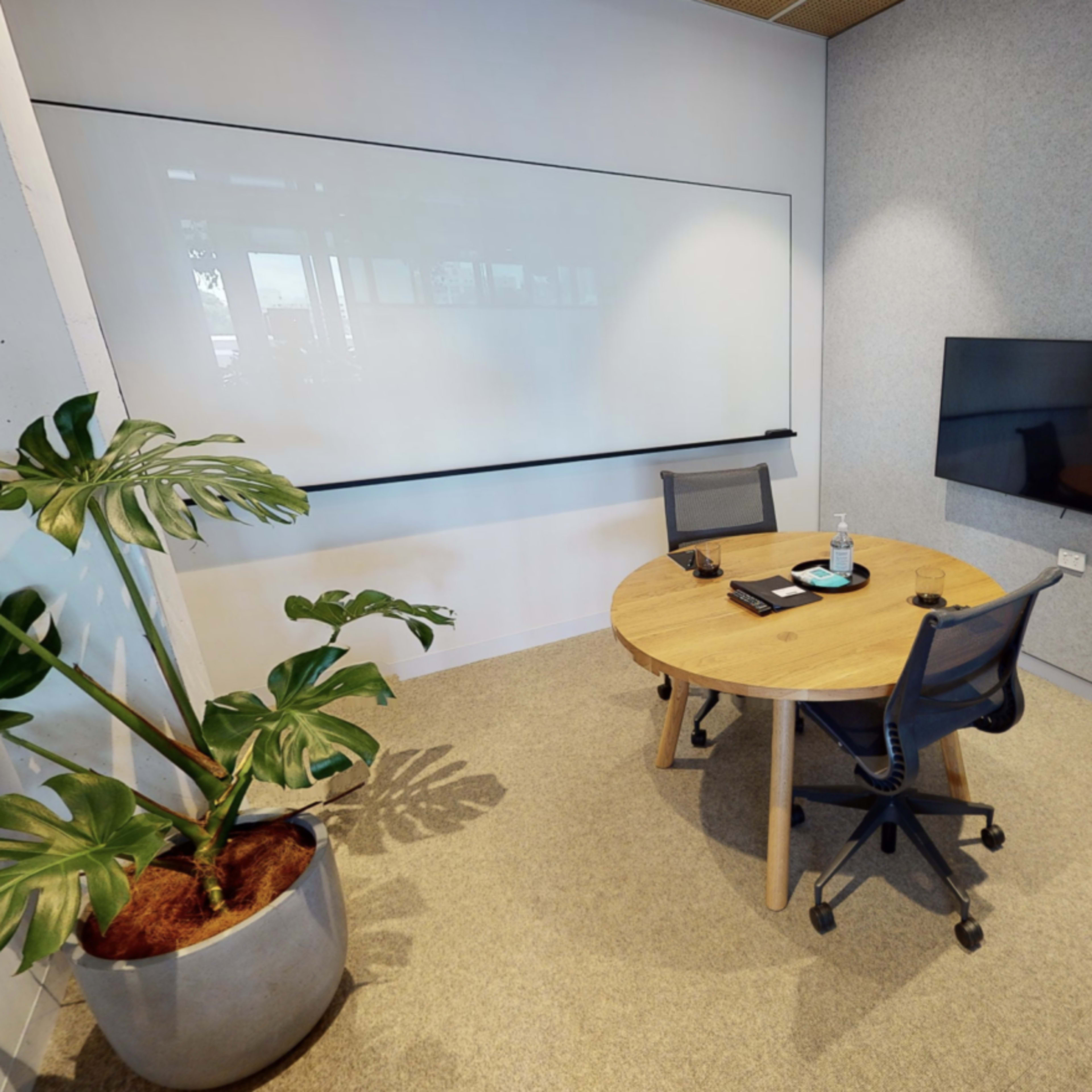 A minimalistic meeting room features a round wooden table with two chairs and a large wall-mounted screen, accompanied by a potted plant in the corner.