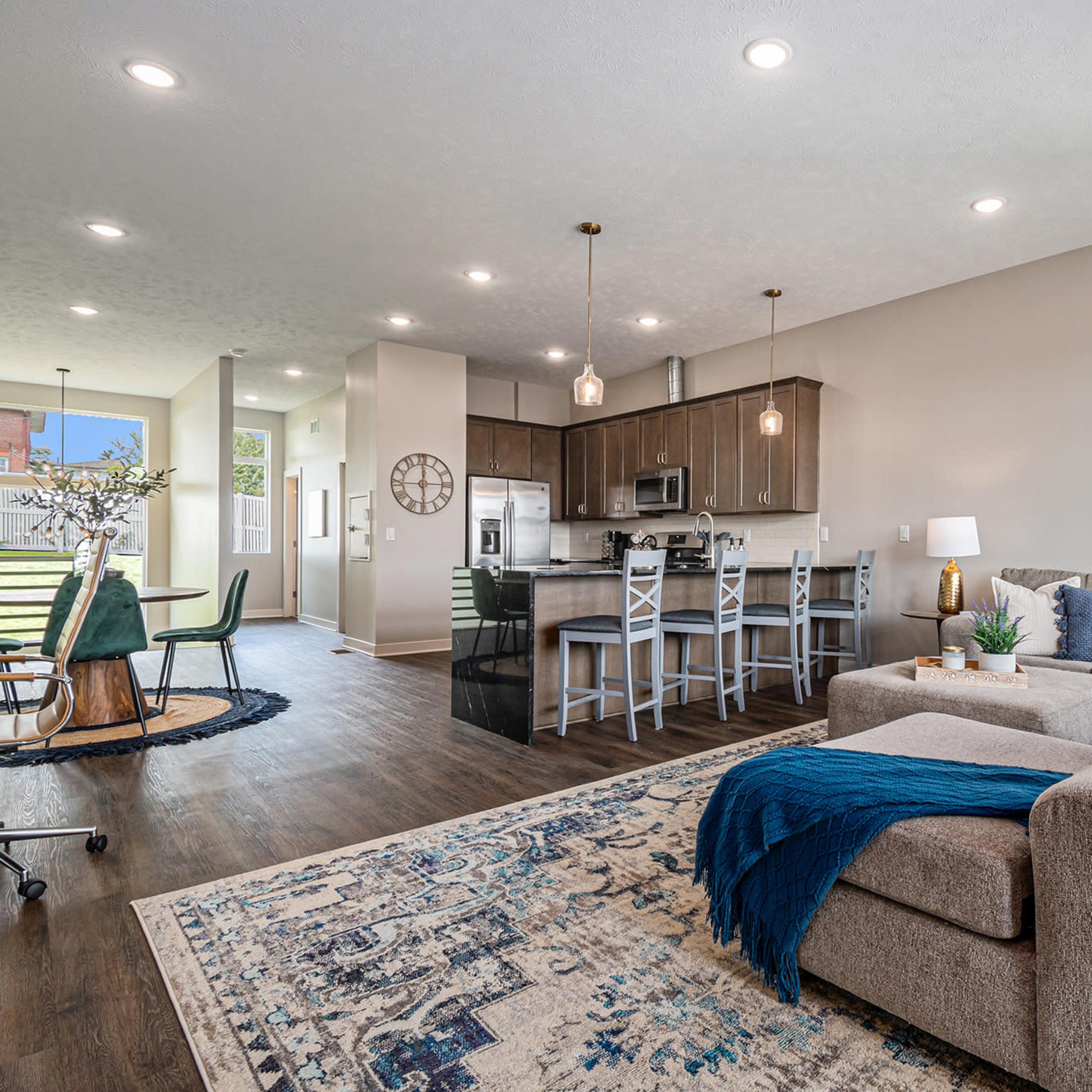 A bright, open-concept living and dining area featuring a modern kitchen with dark cabinetry, a comfortable sofa, and a large area rug.