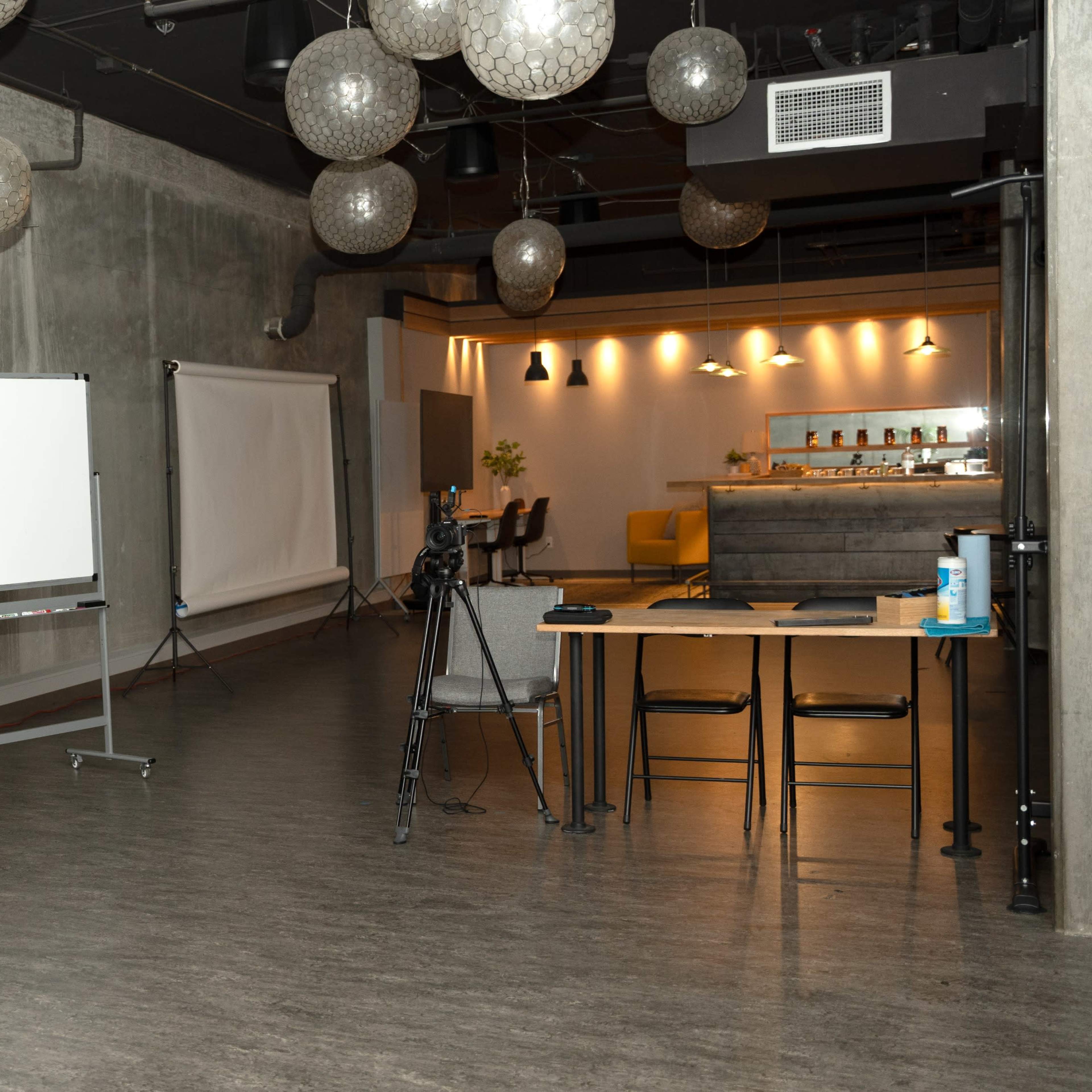 A spacious studio with concrete walls, equipped with lighting equipment, a whiteboard, and a table set up with chairs.