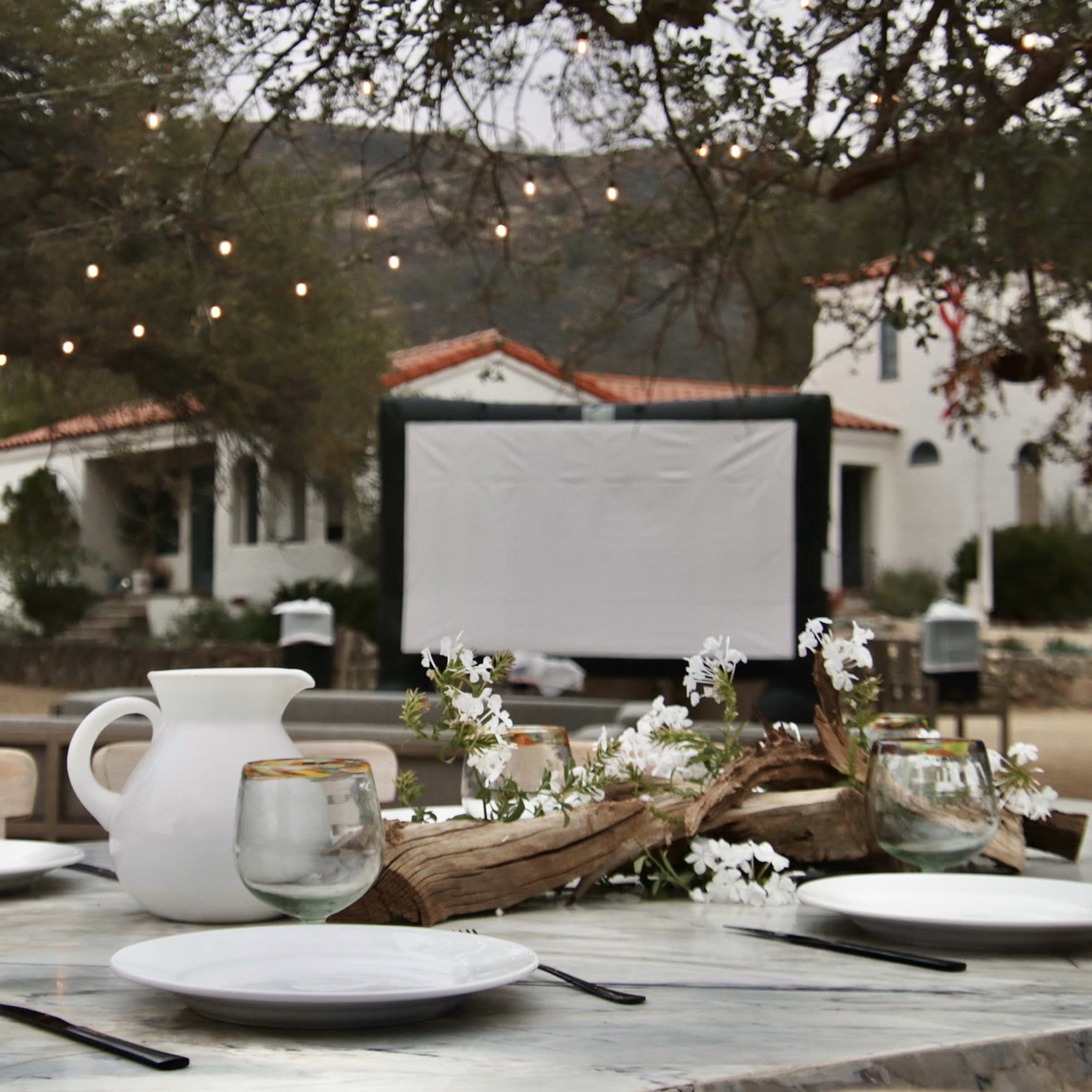 A rustic outdoor dining setup with a table adorned with plates, glasses, and a central arrangement of flowers, set against a backdrop of white buildings and string lights.
