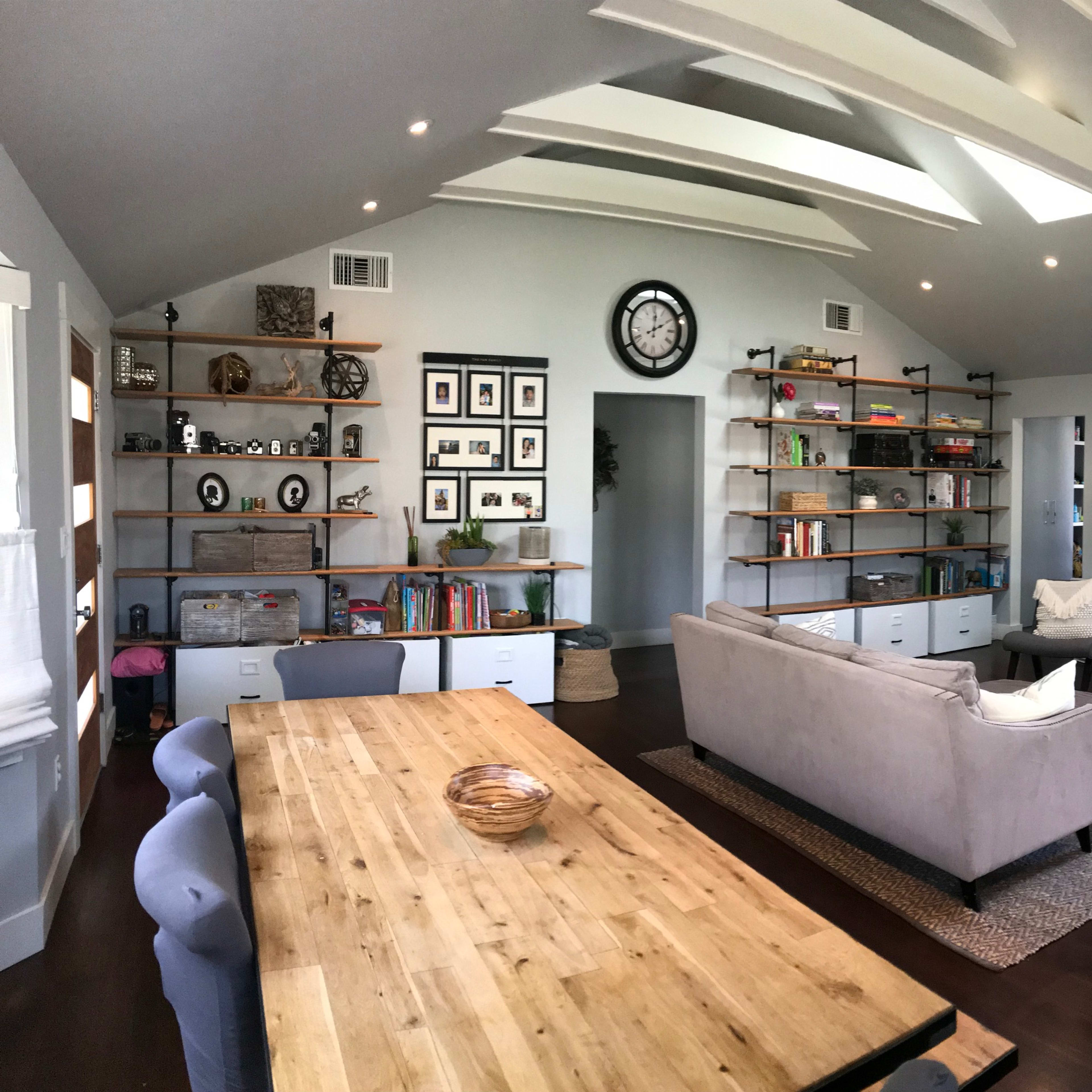 A modern open-concept living area with a kitchen, dining space, and a lounge that features shelves, a clock, and large windows.