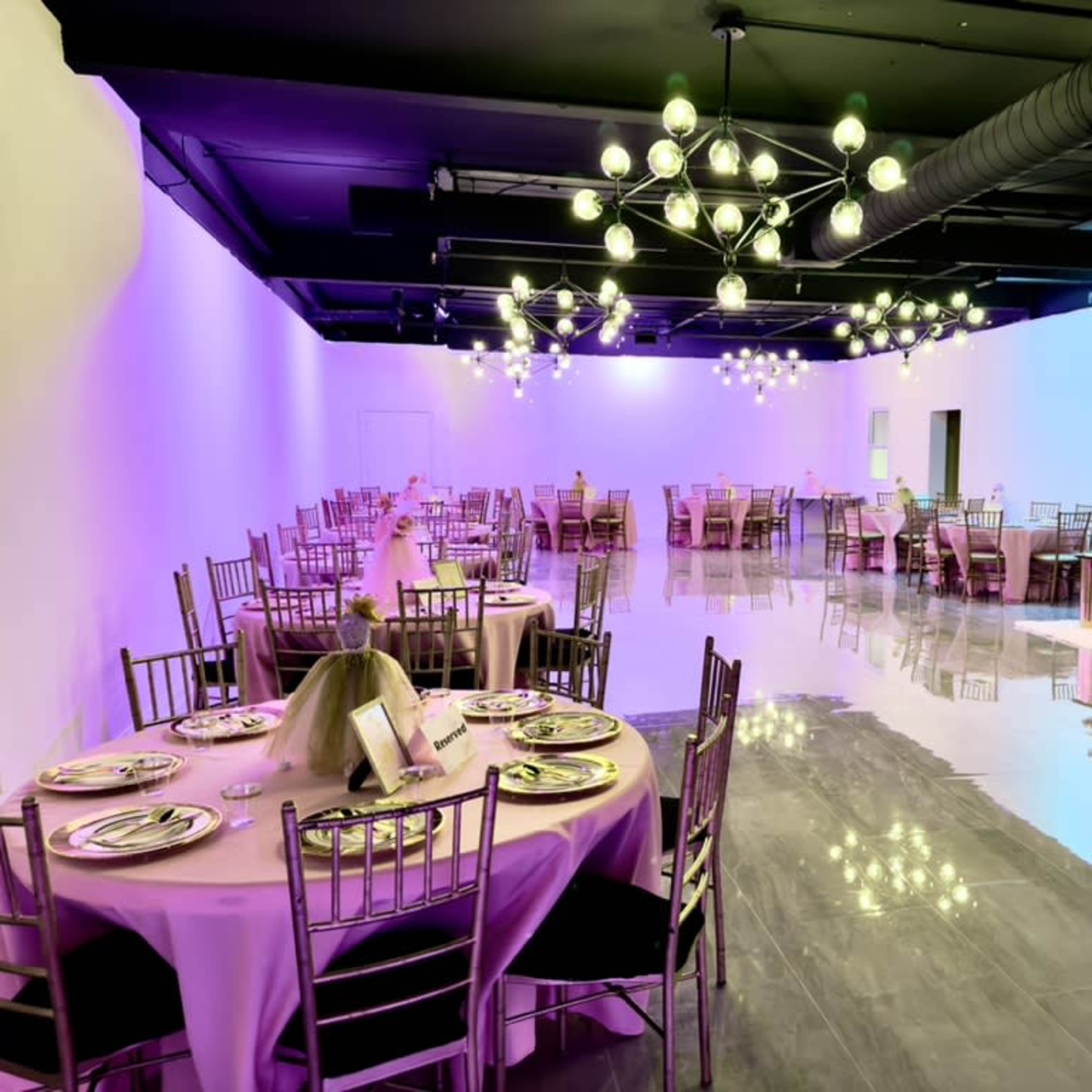 A spacious event hall is set up with neatly arranged round and rectangular tables, each adorned with table settings and decorative centerpieces, under a collection of modern light fixtures.