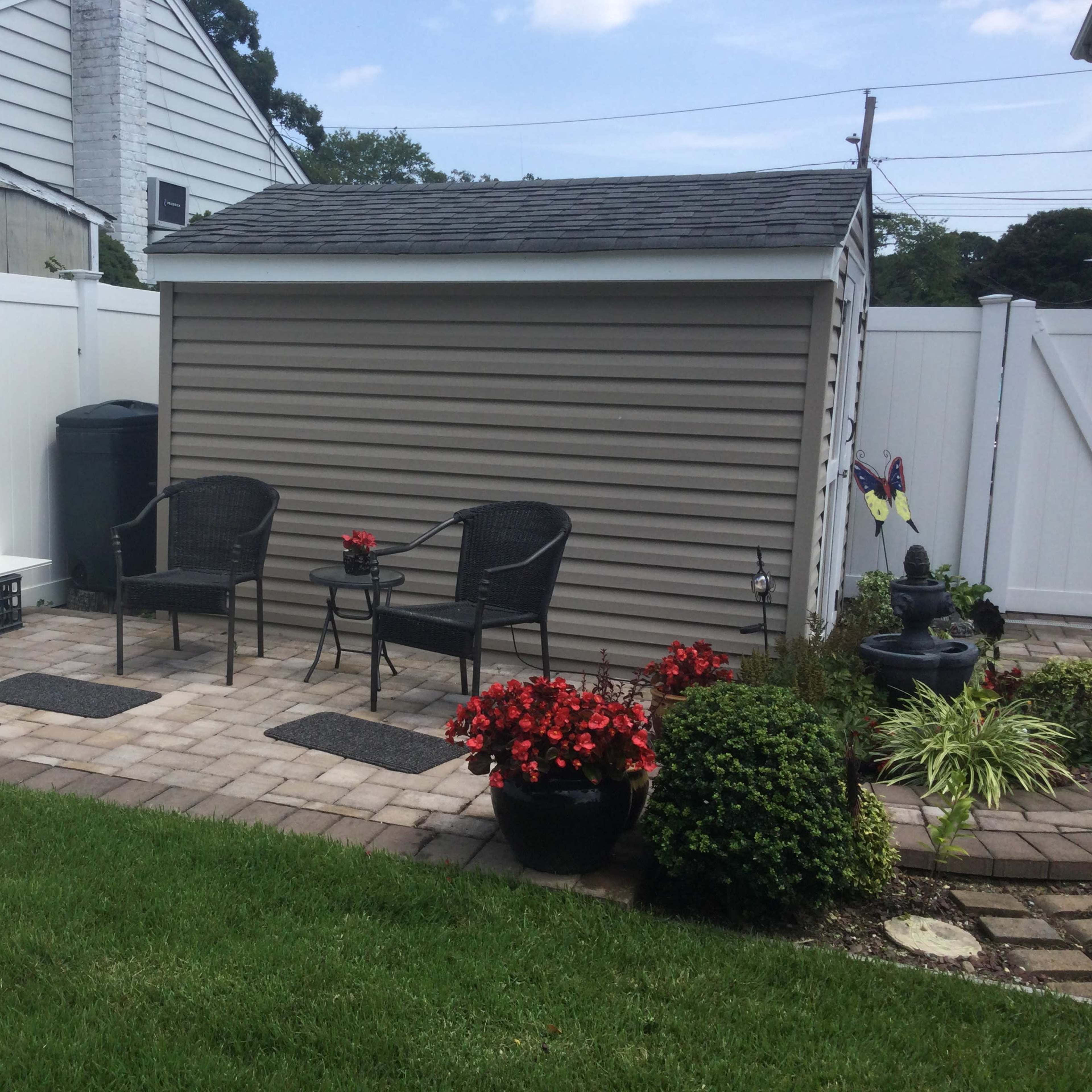 A backyard patio area with two black chairs, a small table, a storage shed, and various potted plants, all surrounded by a white fence.