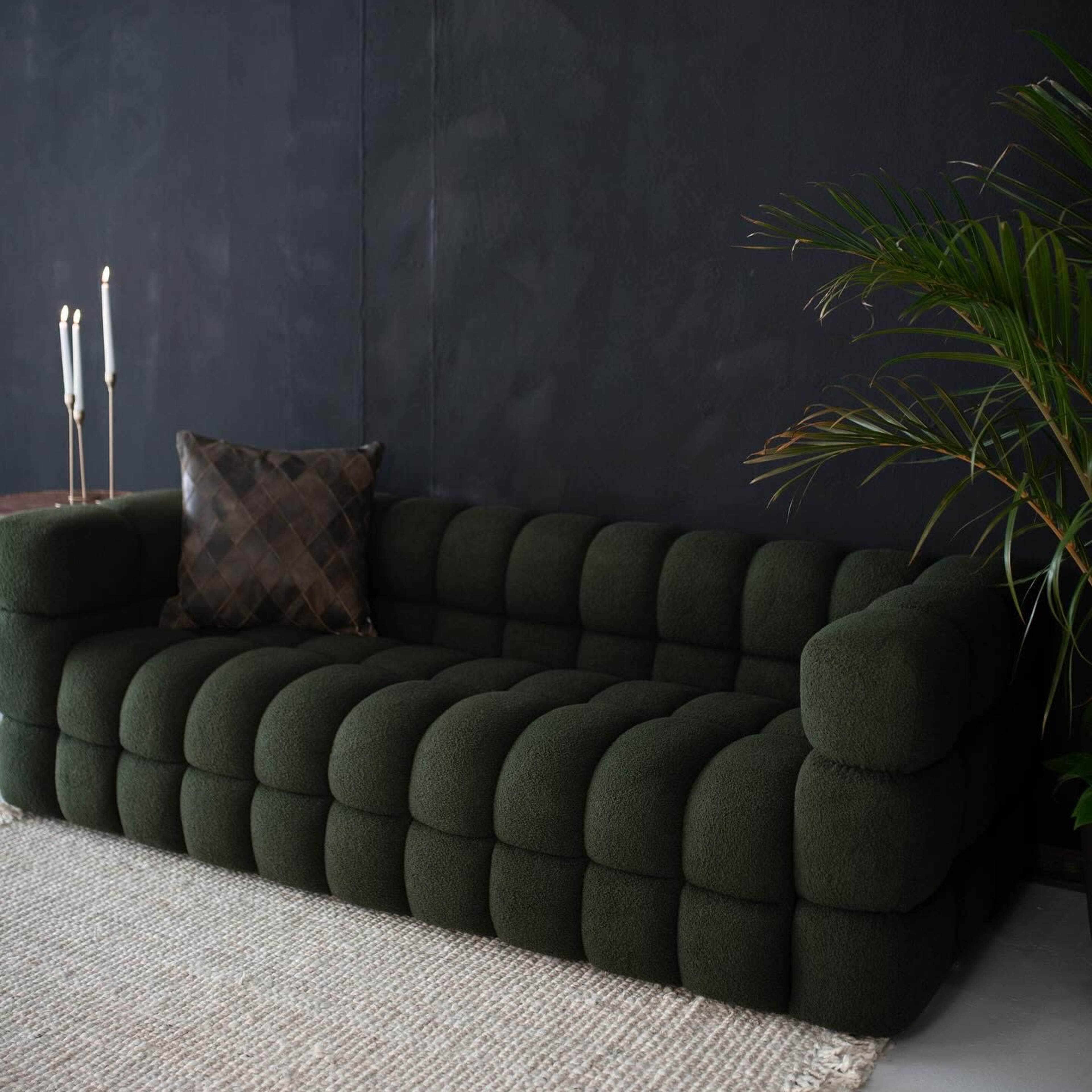 A green, tufted sofa is positioned against a dark wall, accompanied by a decorative pillow and a potted plant nearby.
