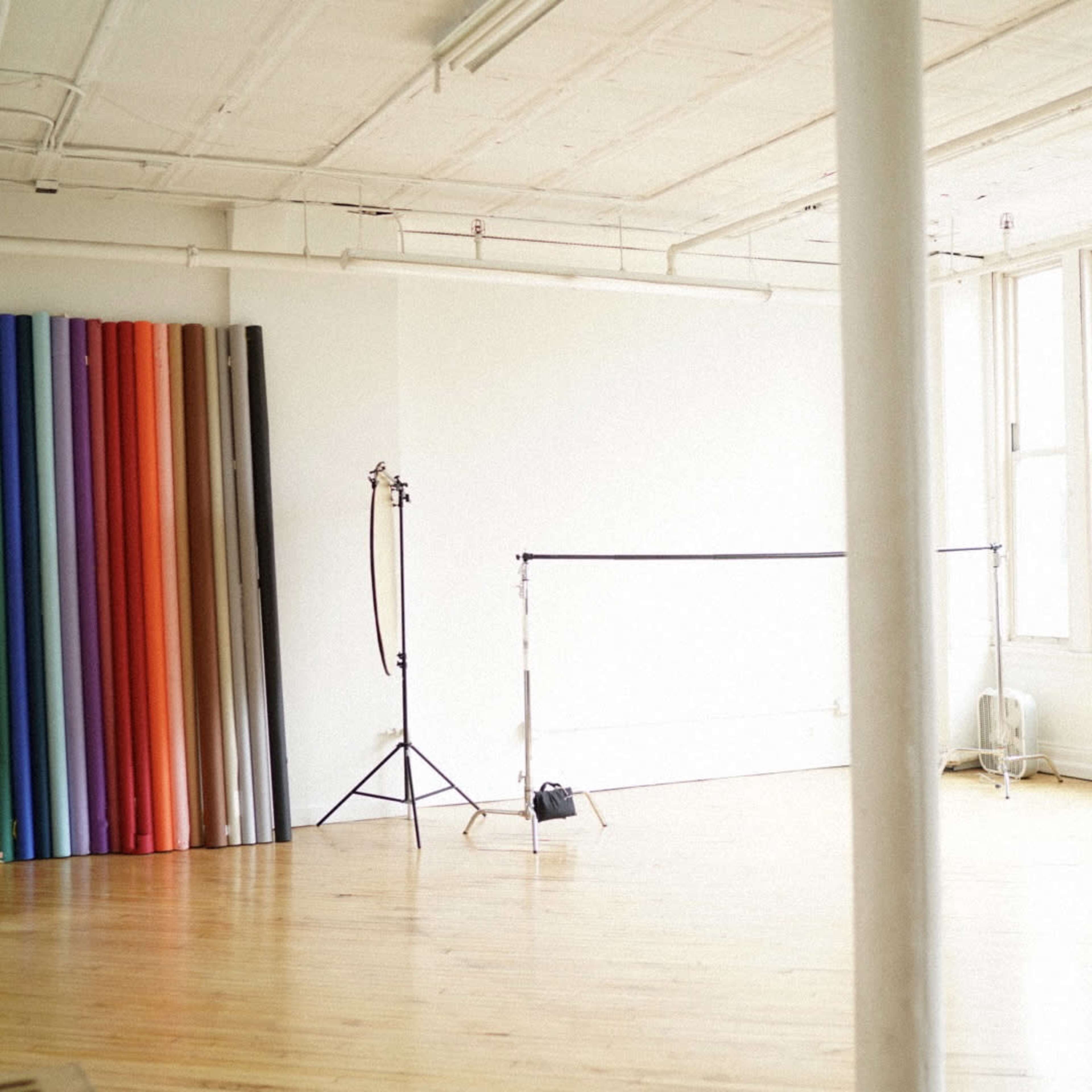 A spacious, empty studio with colorful backdrop rolls, a lighting stand, and a radiator, illuminated by large windows.