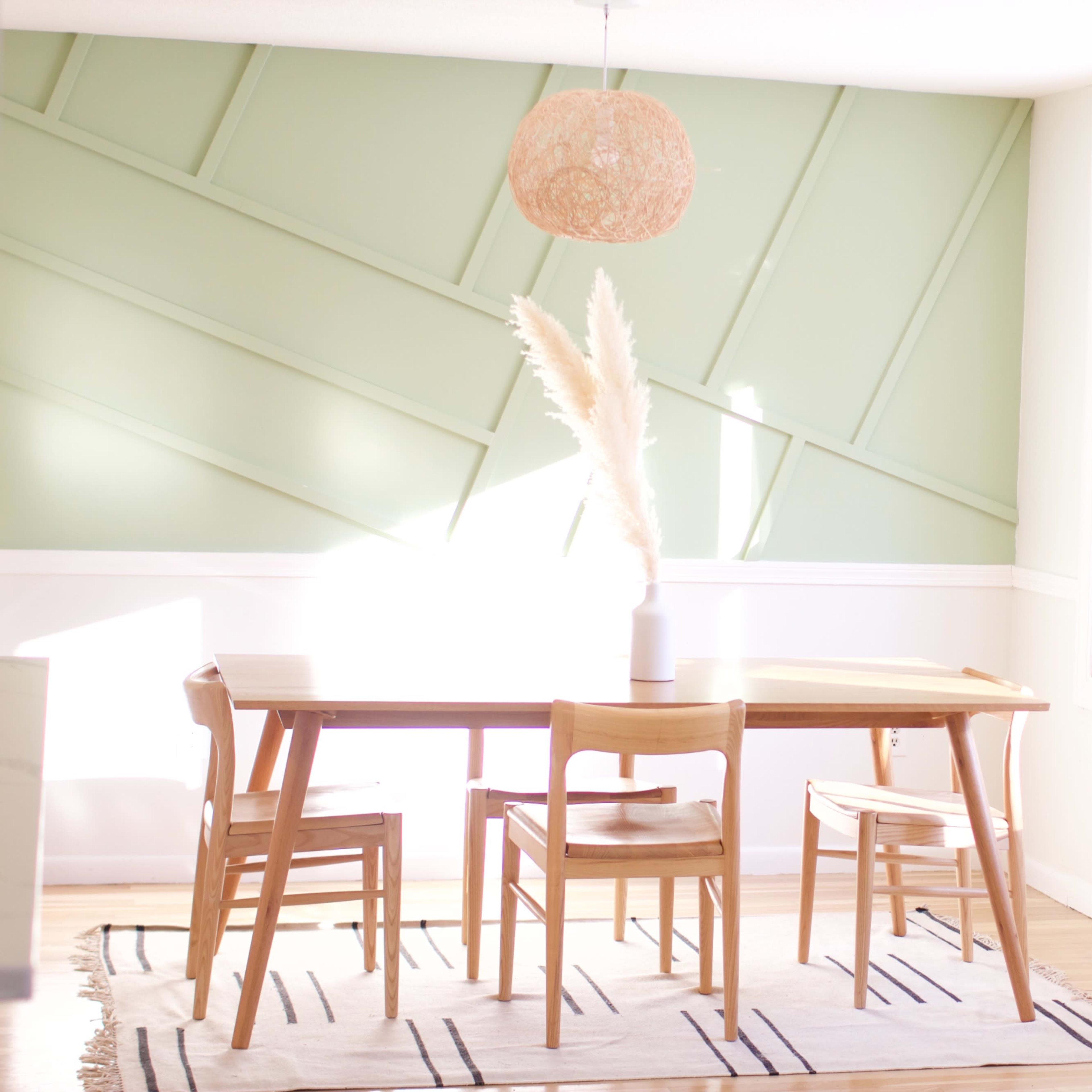 A wooden dining table with four chairs is positioned in a sunlit room featuring a green accent wall and a large window.