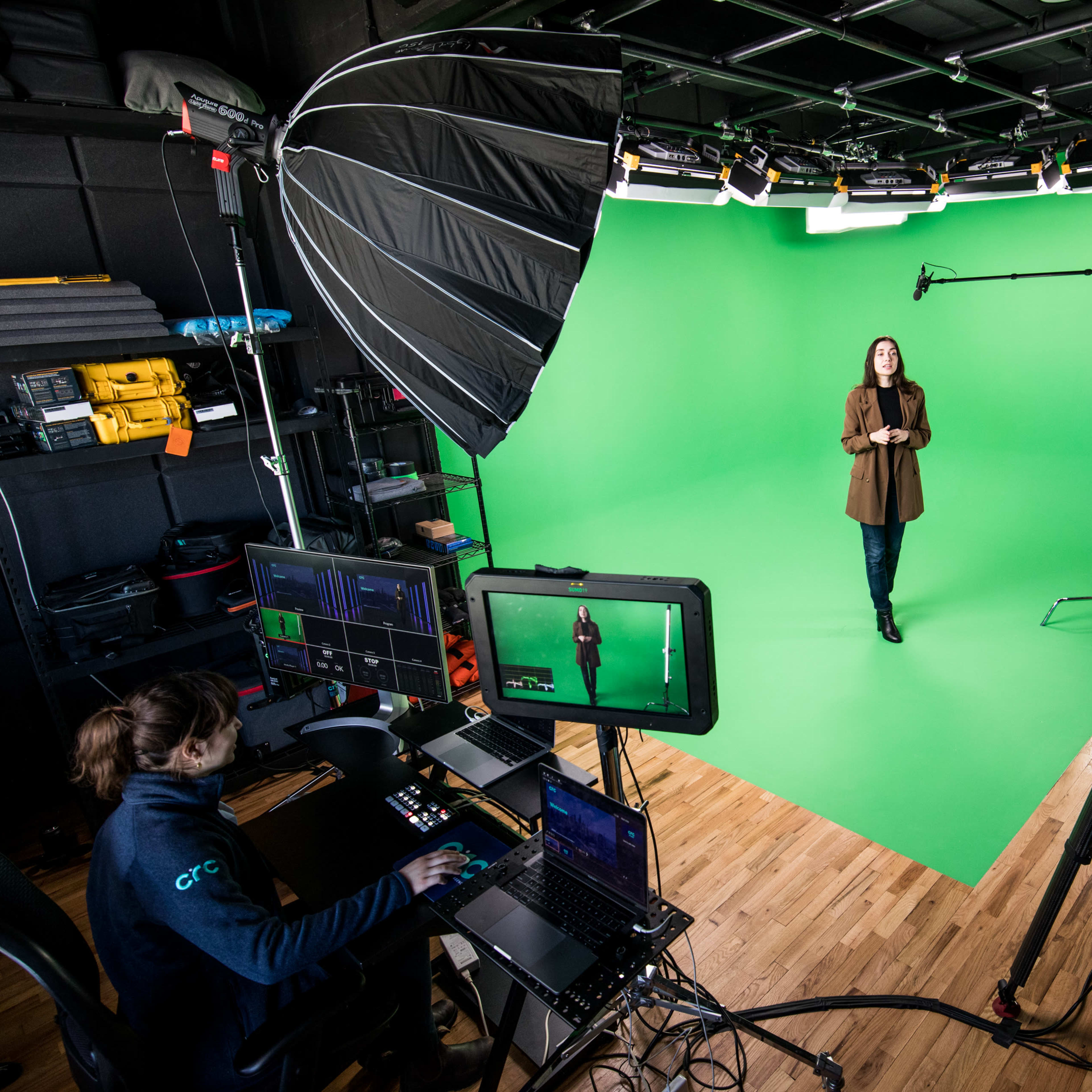 A woman stands in front of a green screen while a person operates equipment at a nearby workstation filled with monitors and cameras.