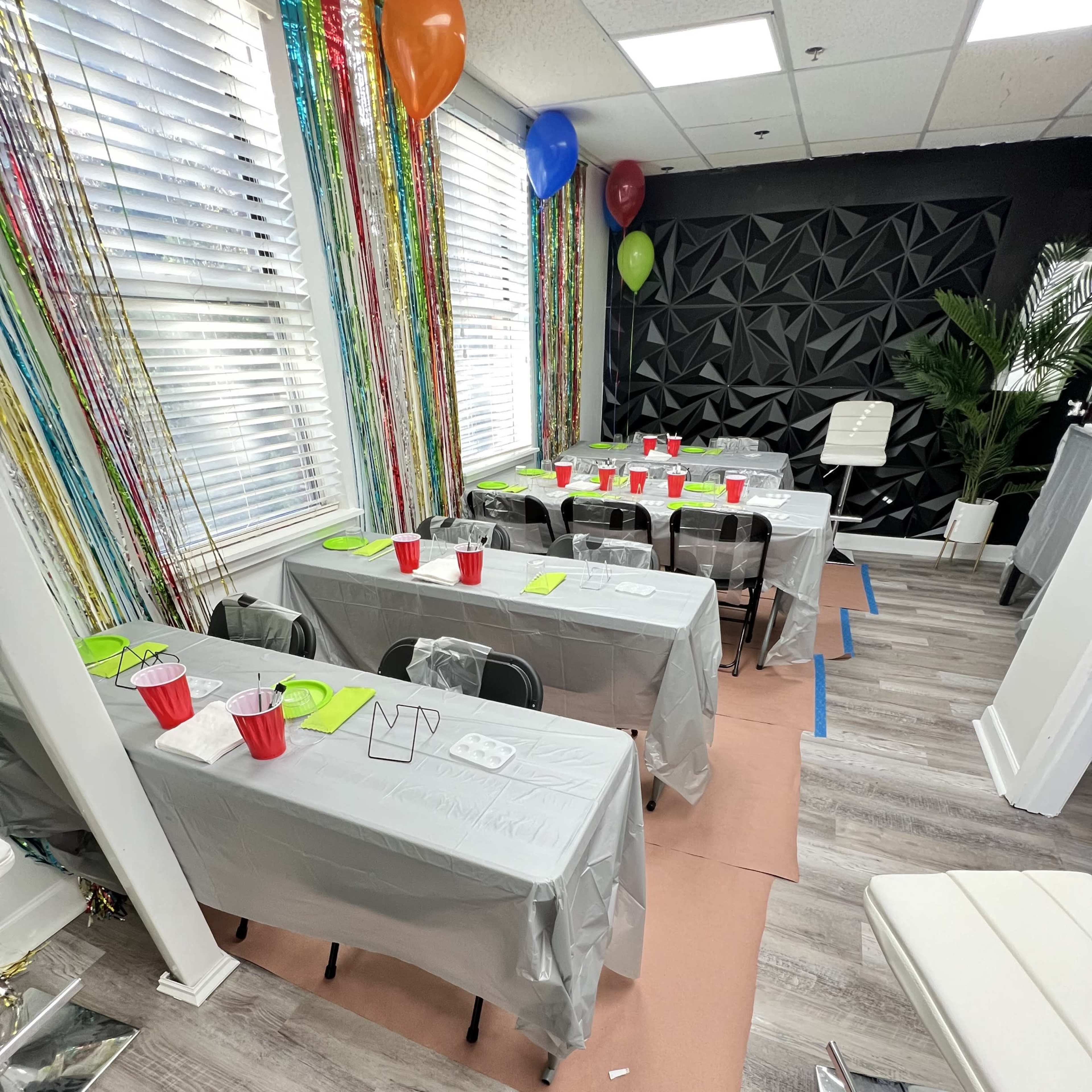 A decorated room with several tables covered in gray tablecloths, each set with red cups and green placemats, surrounded by colorful streamers and balloons.