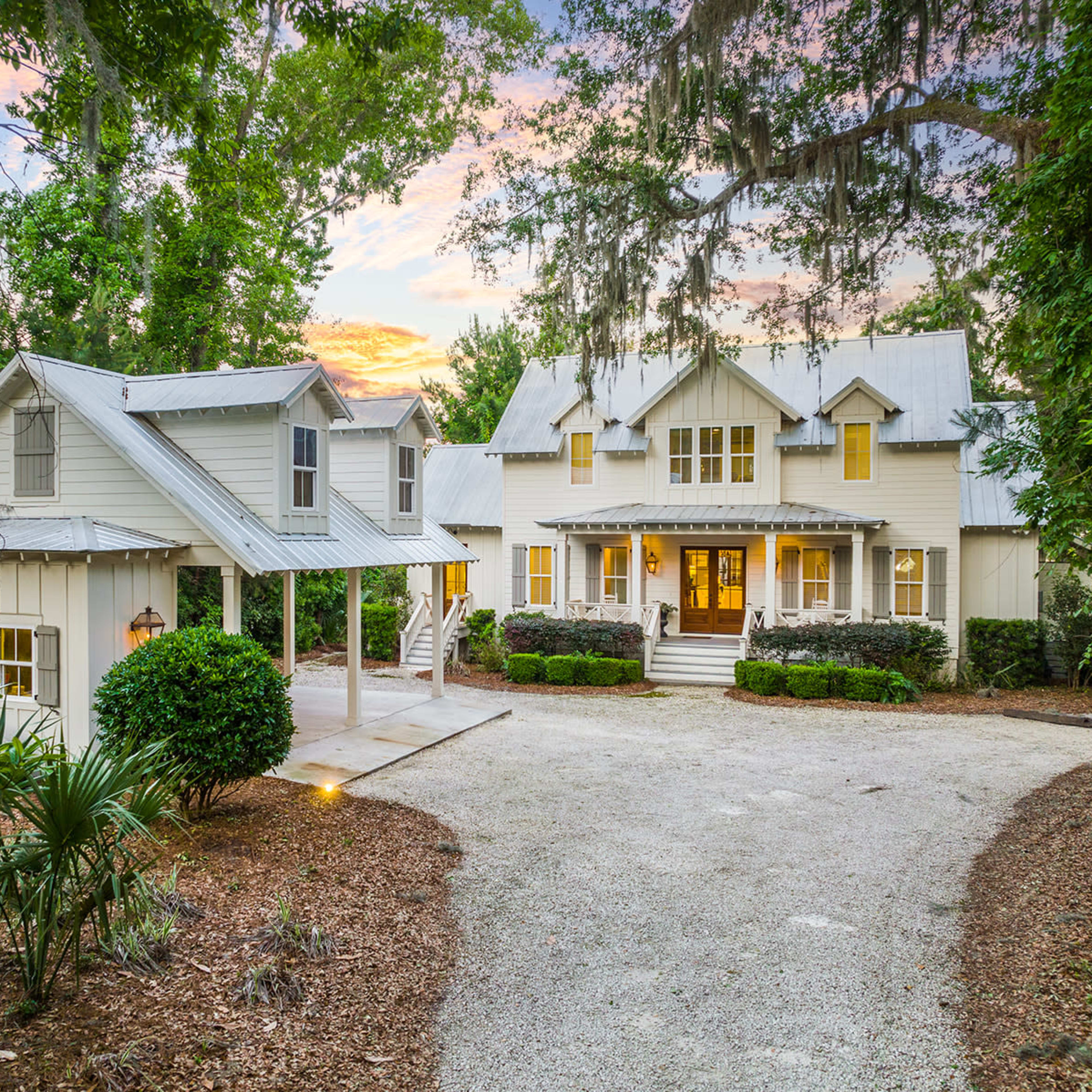 A white, two-story house with a front porch and multiple gabled roofs is set among trees and landscaped greenery in a gravel driveway.
