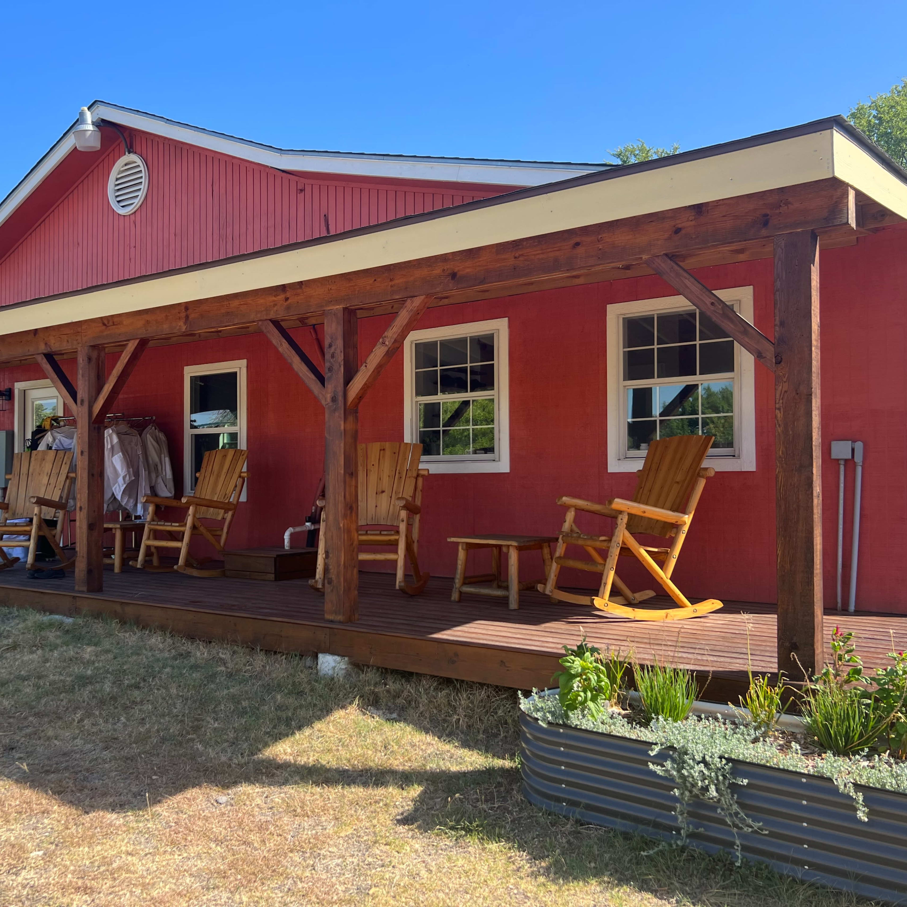 A red house with a covered porch features several wooden rocking chairs and a few plants in a garden bed.