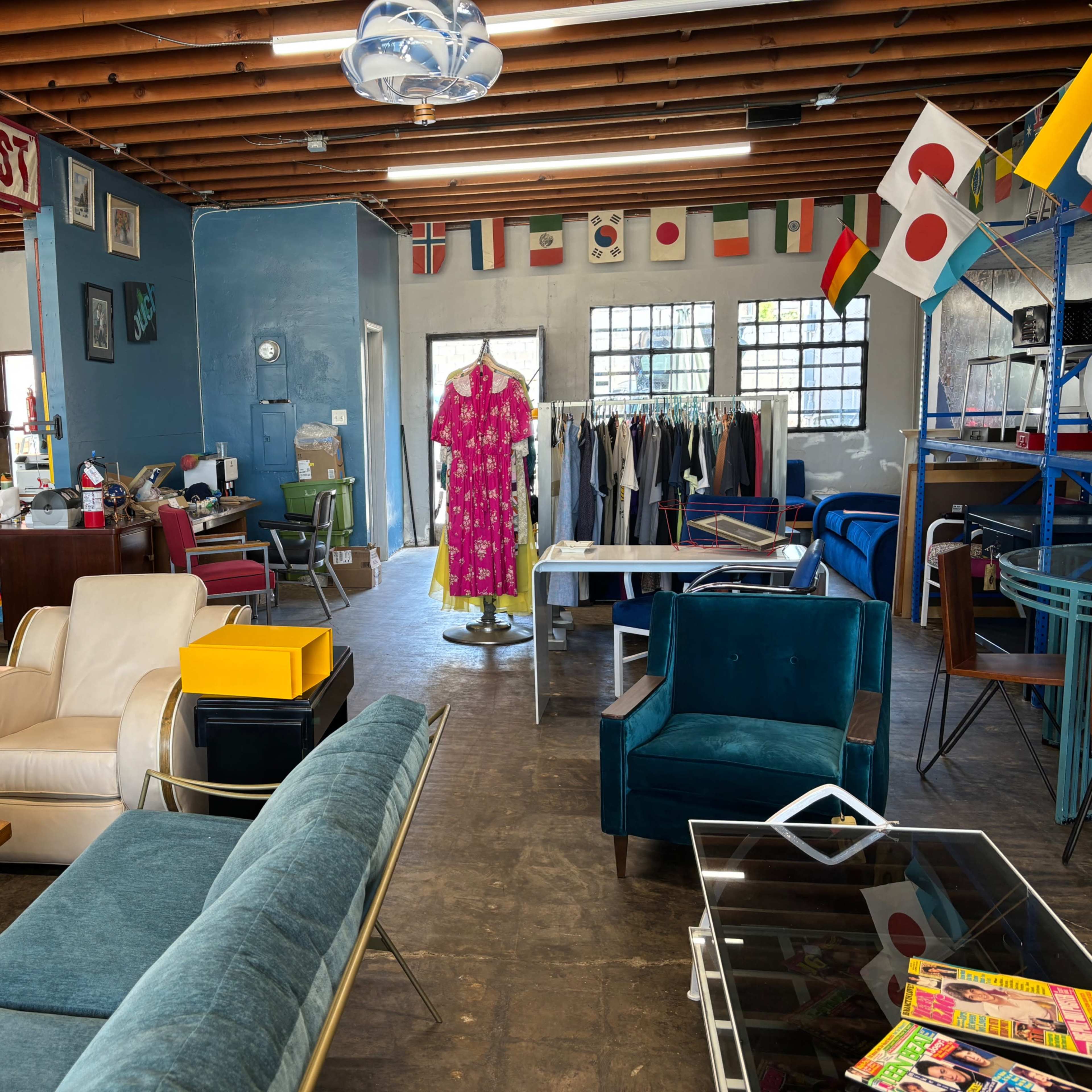 A brightly lit thrift store with various pieces of furniture, including chairs, a sofa, and clothing racks adorned with different national flags.