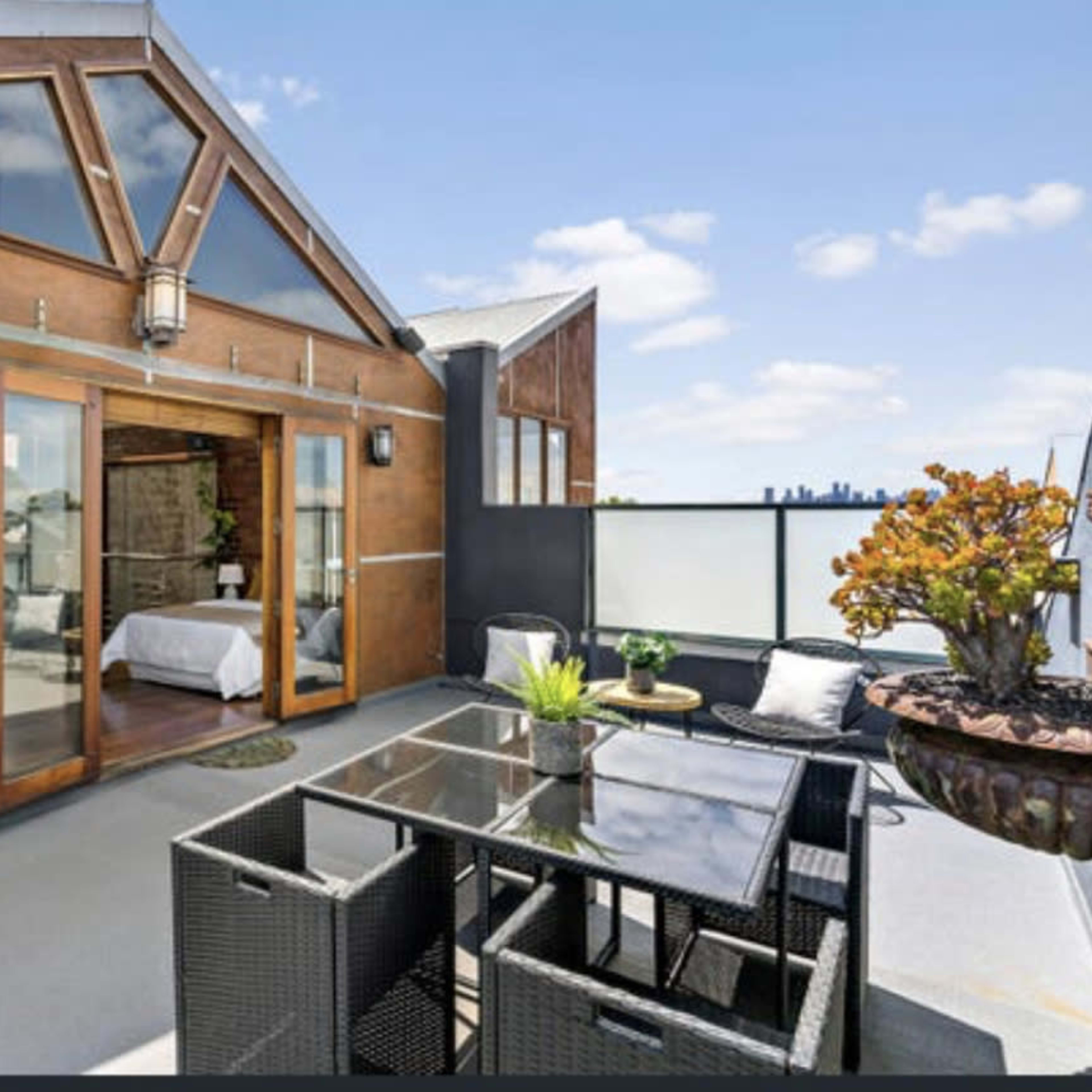 A modern rooftop patio featuring a glass table surrounded by black wicker chairs, with sliding glass doors leading into an interior space.