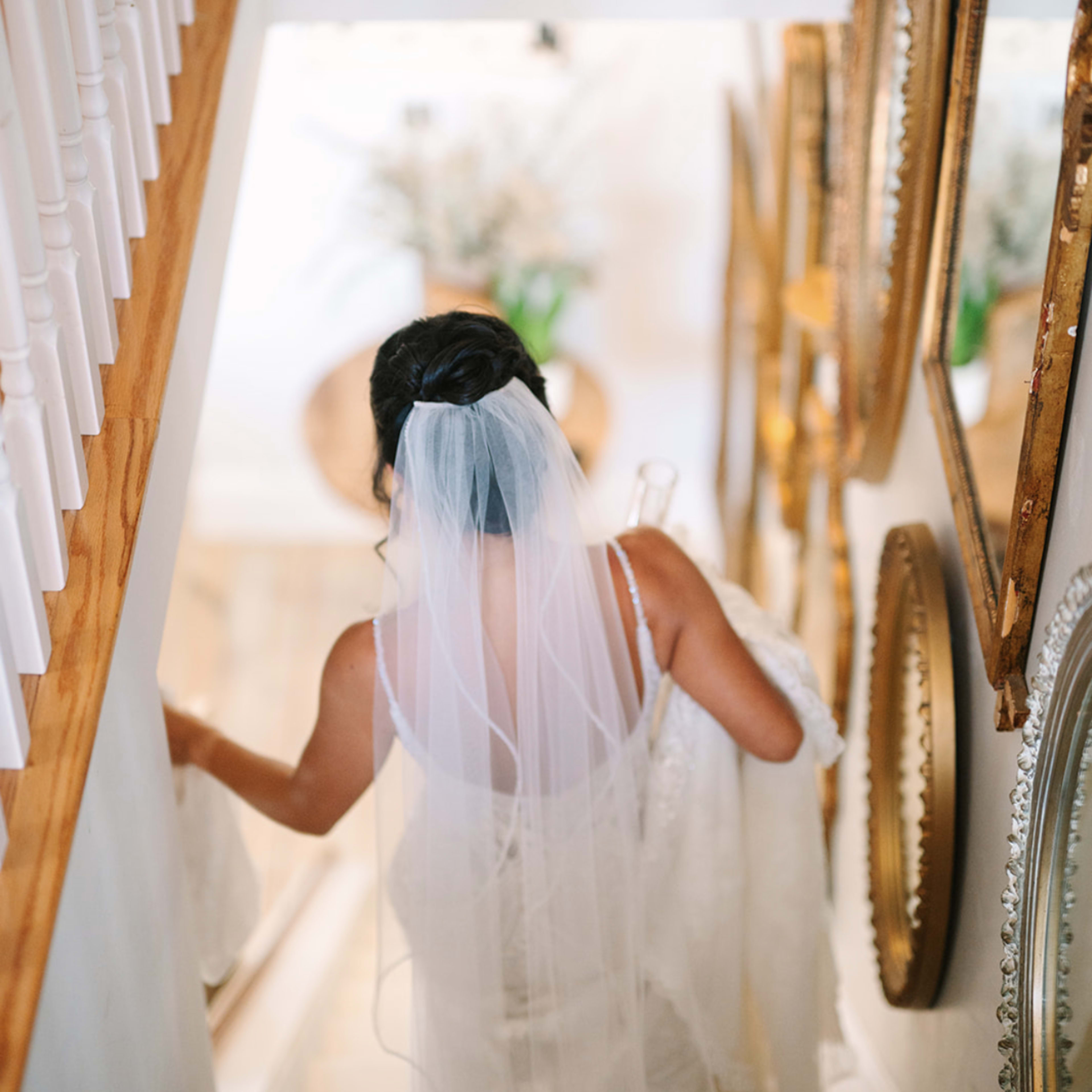 A woman in a wedding dress and veil ascends a staircase adorned with framed mirrors.
