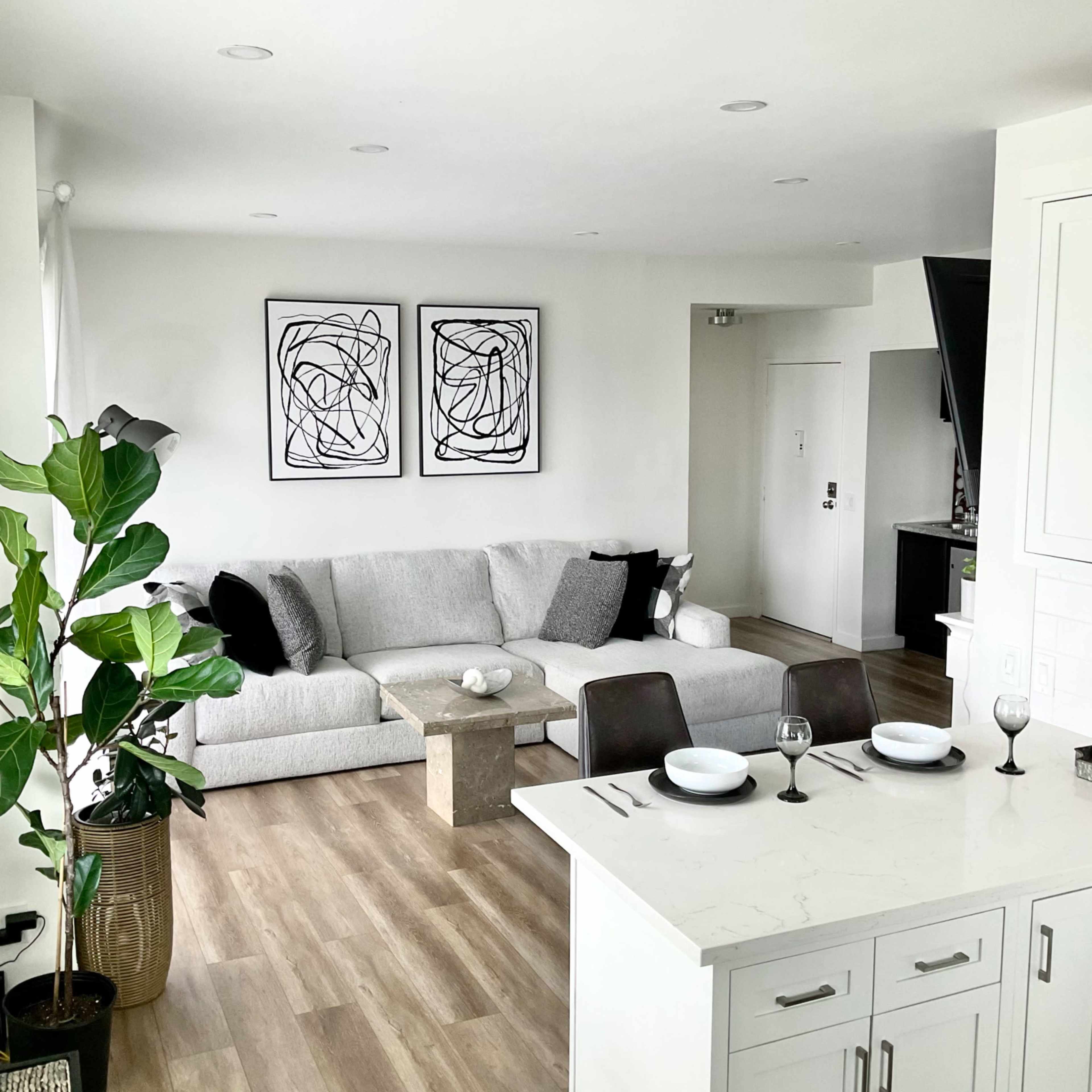 A modern living room featuring a light gray sectional sofa, two abstract black and white framed prints on the wall, and a dining area with a light-colored countertop and table settings.