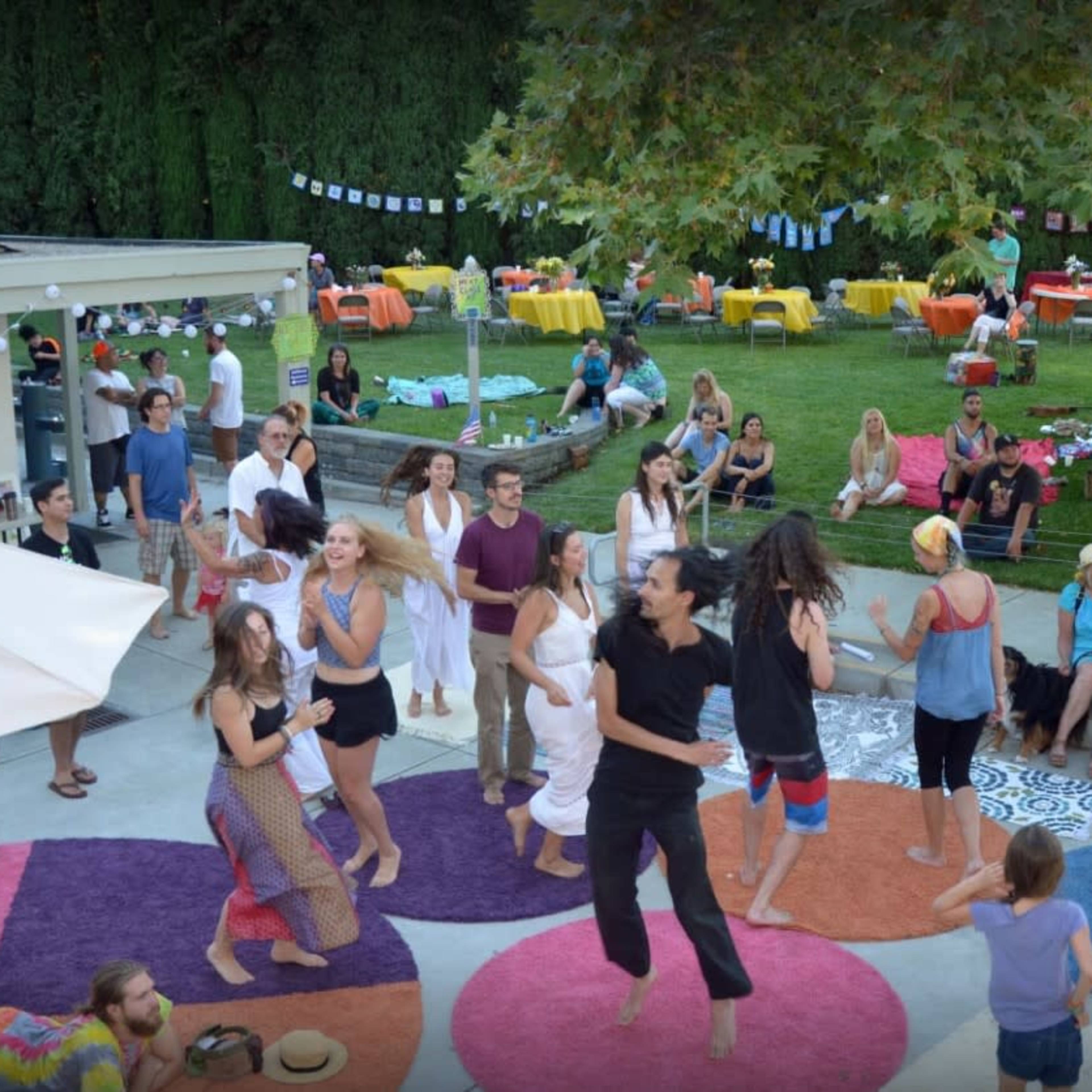 A group of people is dancing on colorful rugs in a backyard party setting, with tables and seating arranged in the background.
