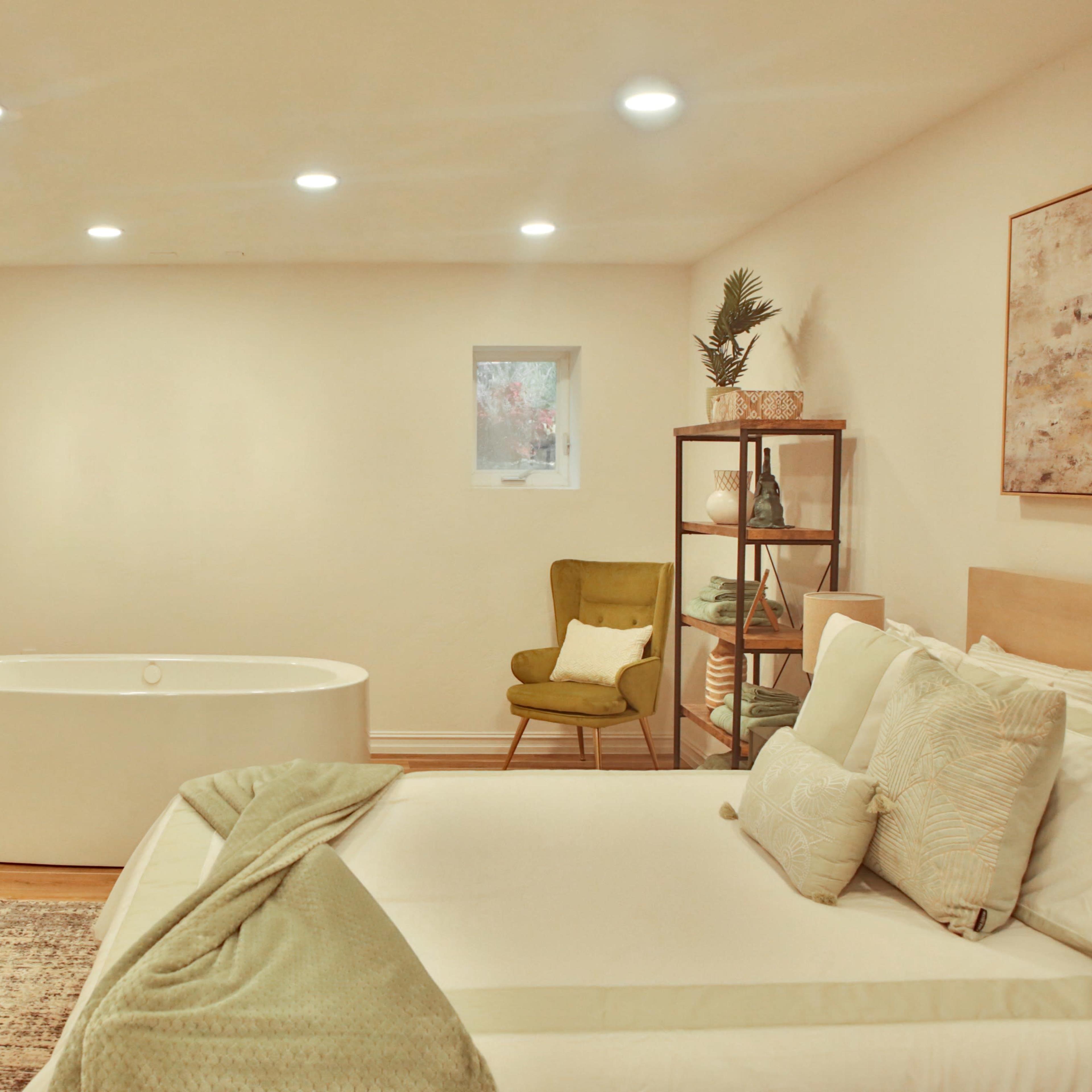 A modern bedroom featuring a freestanding bathtub, a bed with light bedding, and a small bookshelf in a neutral-toned room.