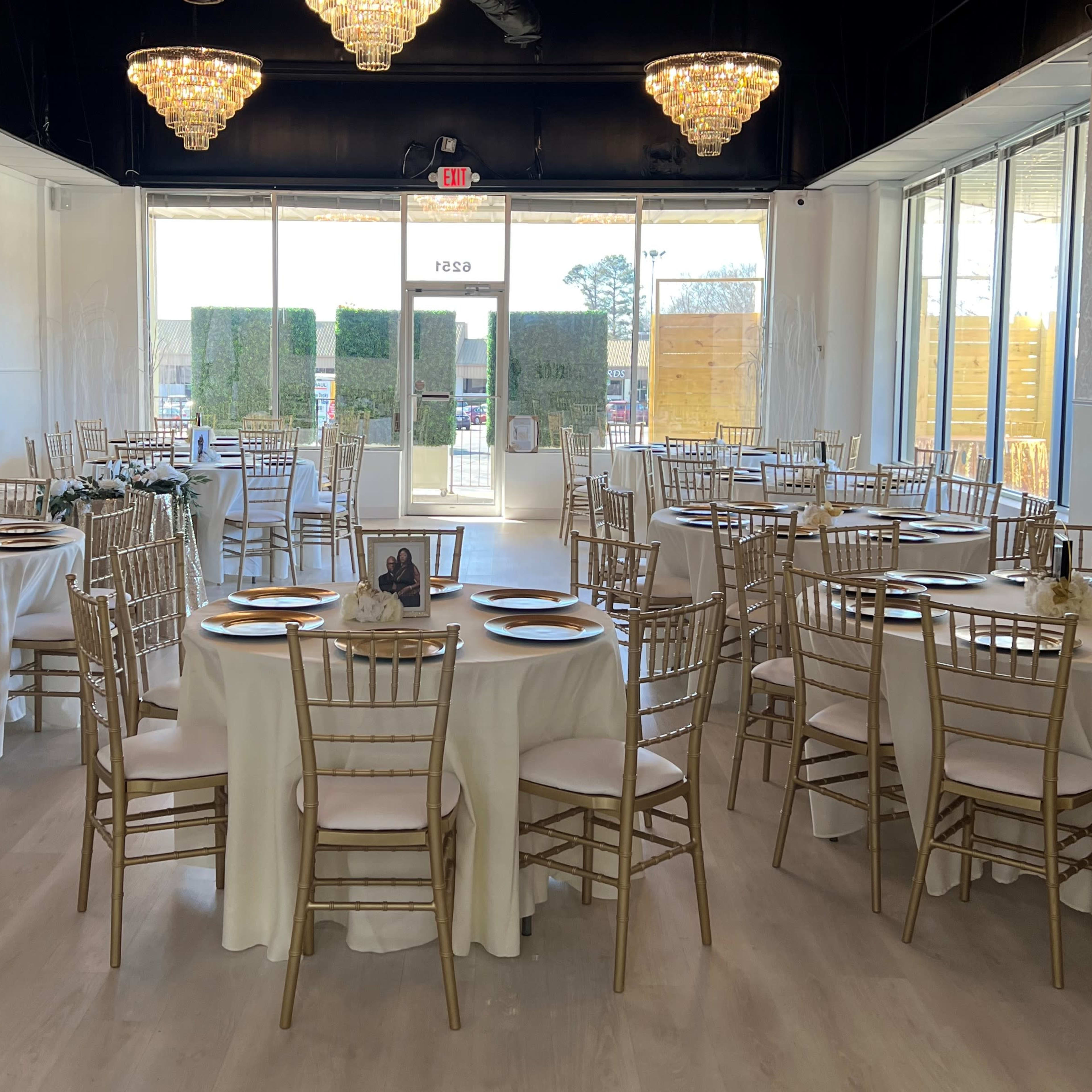 A dining hall set with round tables covered in white tablecloths and adorned with golden chairs, illuminated by chandeliers overhead.