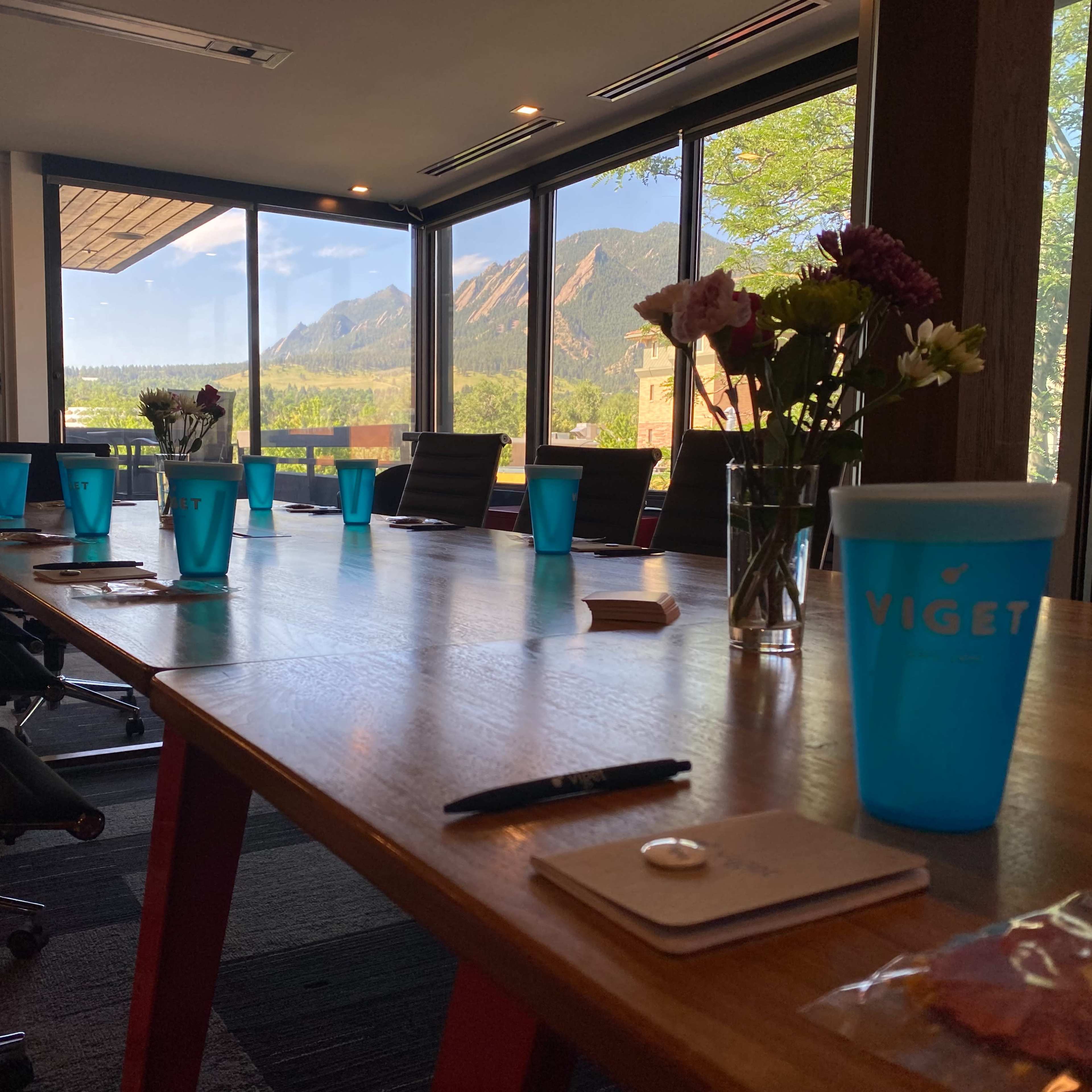 A conference room features a long wooden table set with blue cups, notepads, and a pen, overlooking a scenic view of mountains through large windows.