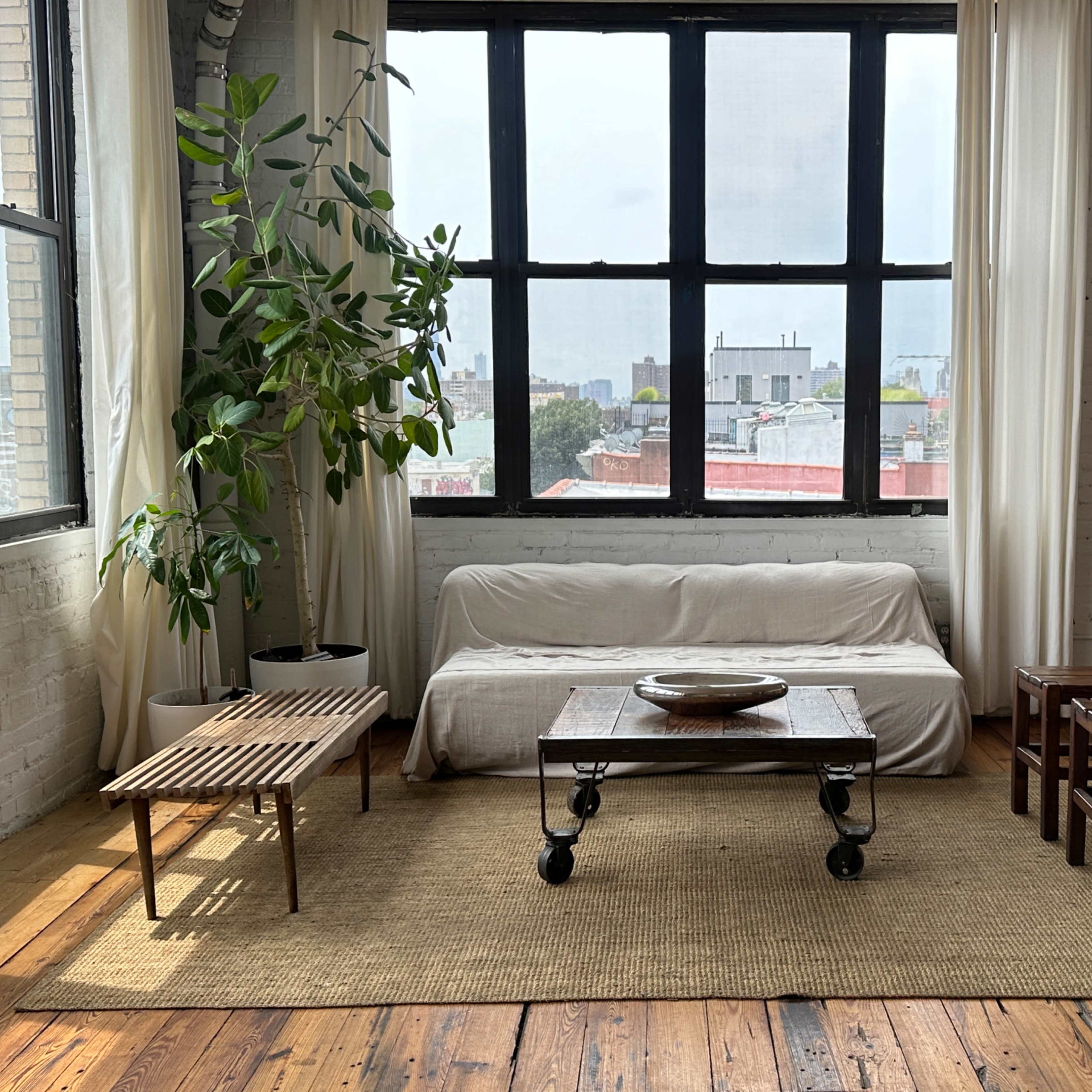 A spacious room features a white sofa, a coffee table on wheels, wooden stools, and a tall plant near large windows with white curtains.