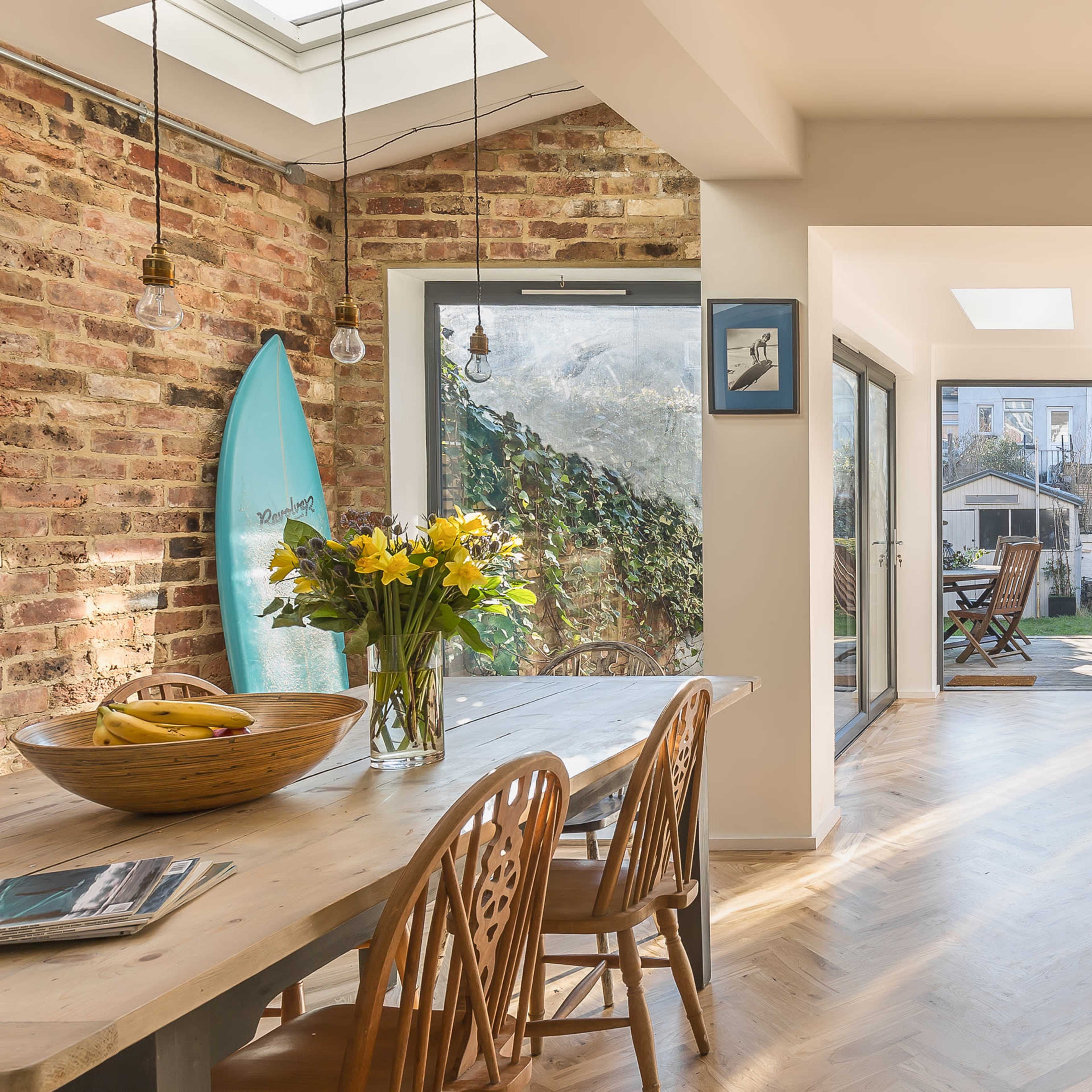 A modern kitchen and dining area features a wooden table with chairs, a bouquet of flowers, and large glass doors opening to a sunny garden with a surfboard leaning against the wall.