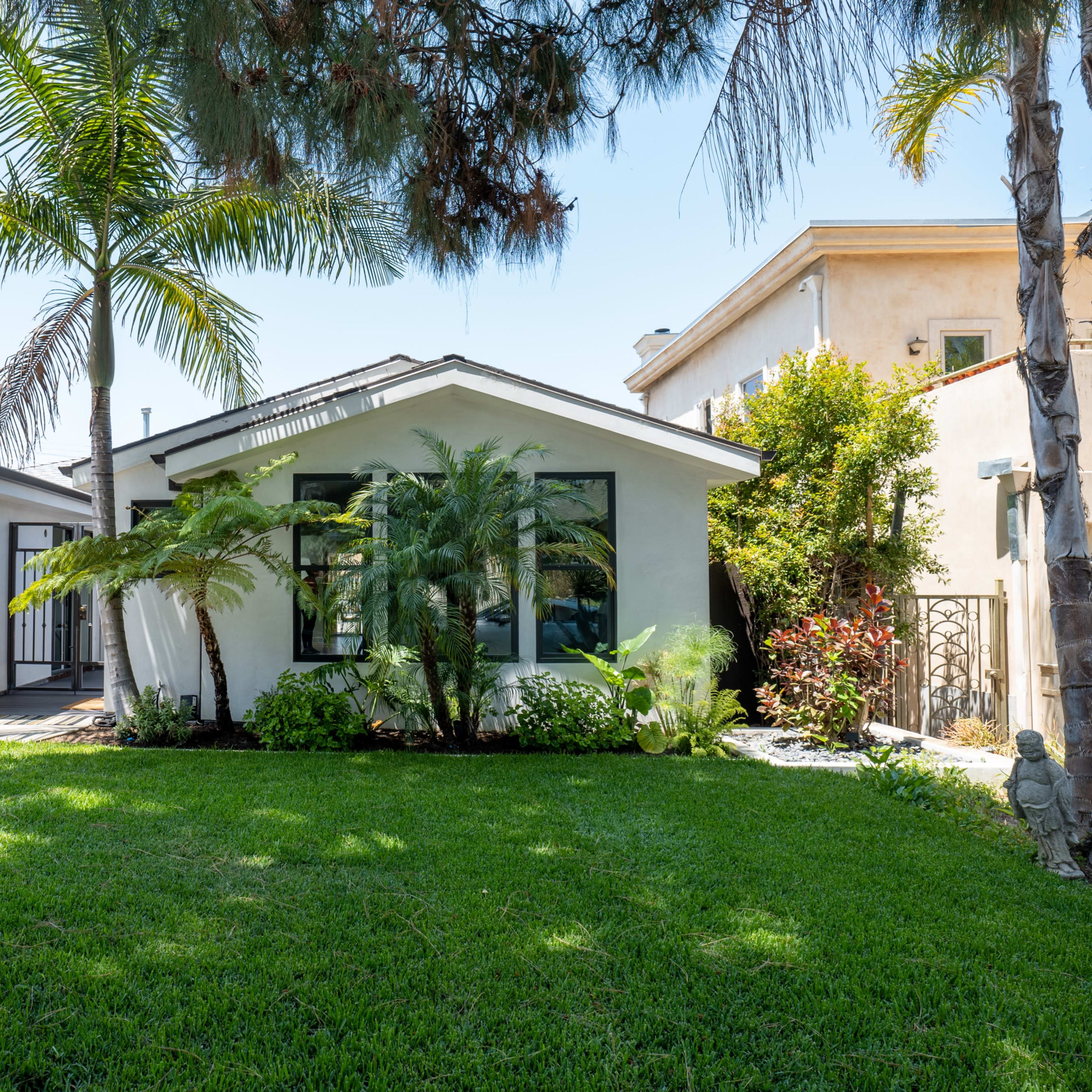 A single-story house with a manicured lawn, palm trees, and tropical plants is shown beside a garage with a yellow exterior.
