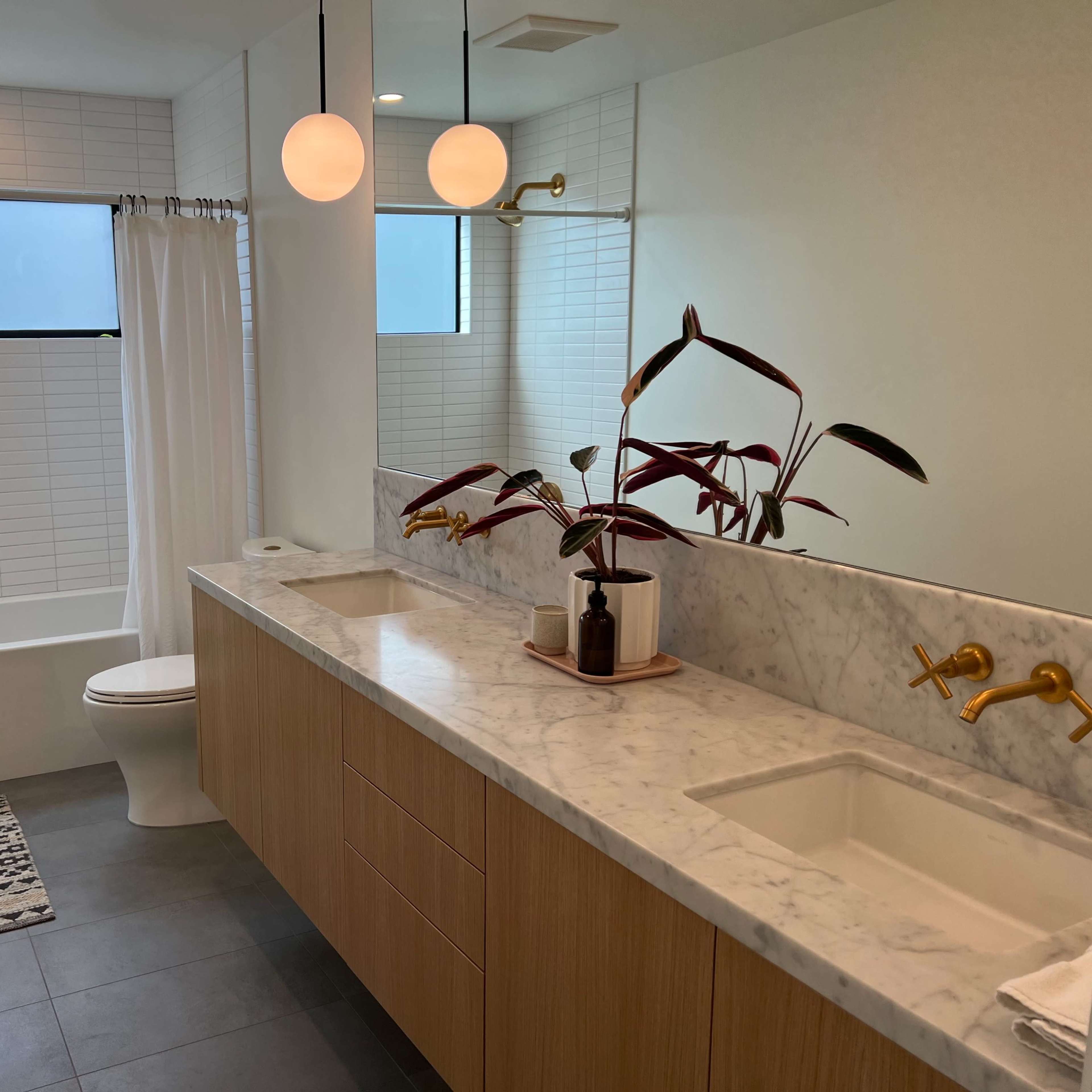 A modern bathroom with a double sink vanity, a large mirror, and a bathtub beside a window, featuring light-colored walls and a plant on the counter.