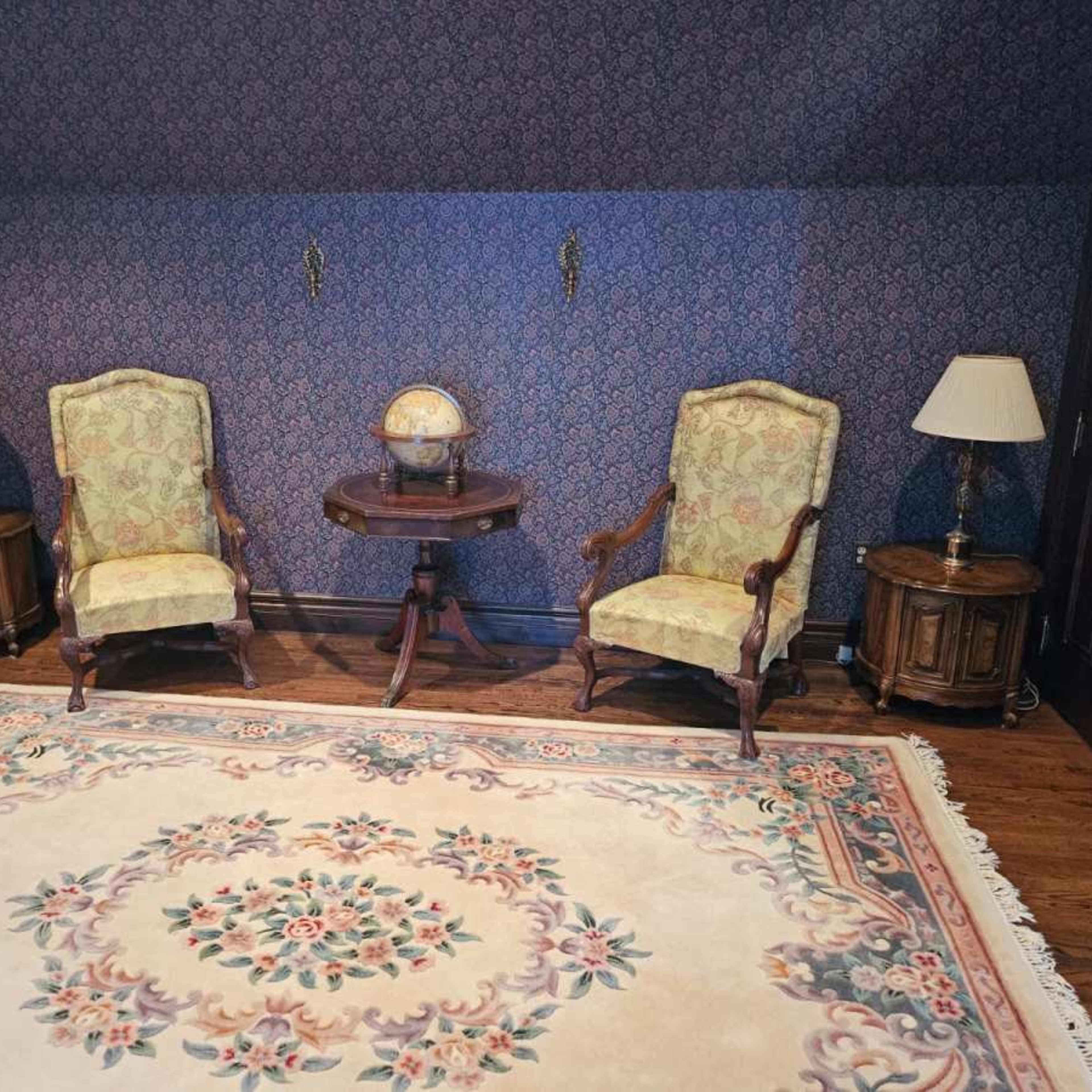 A room with two upholstered chairs, a small round table, a globe, two lamps, and a decorative rug on wooden flooring.