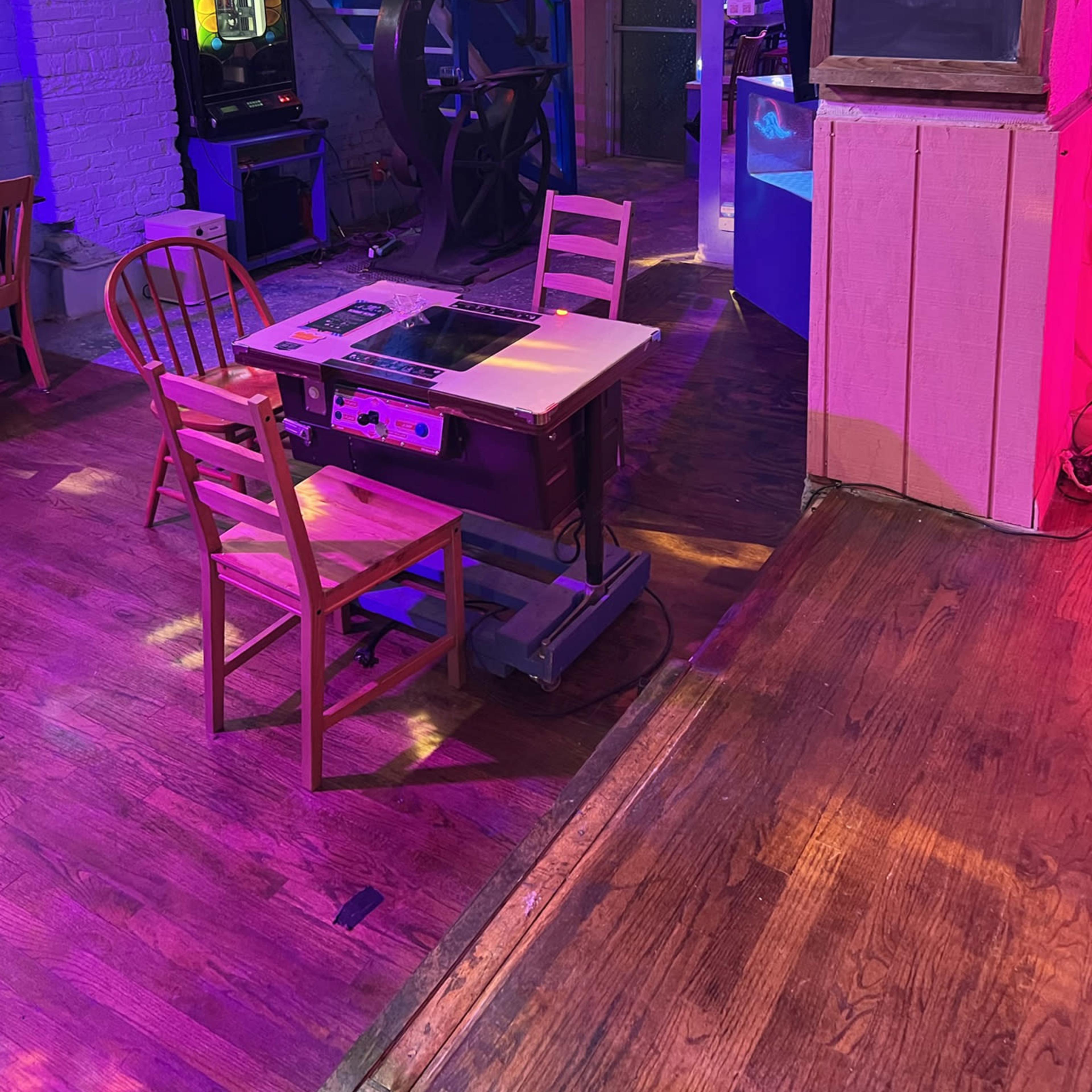 The scene depicts a dimly lit room featuring a vintage table and two wooden chairs near a spiral staircase, with colorful lighting and various retro decor elements.