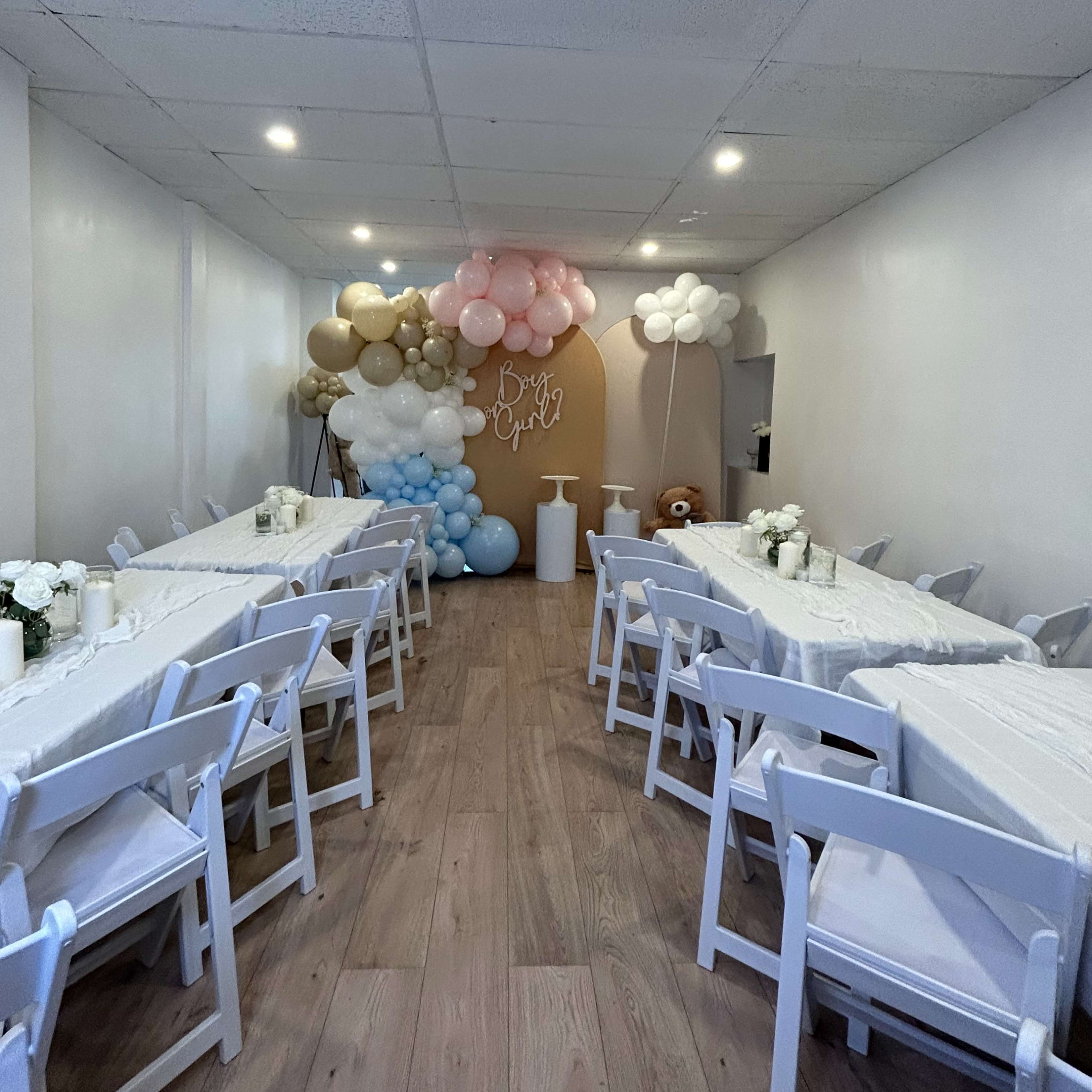 A banquet hall is arranged with white tables and chairs, decorated with balloons in pastel colors, creating a festive atmosphere.