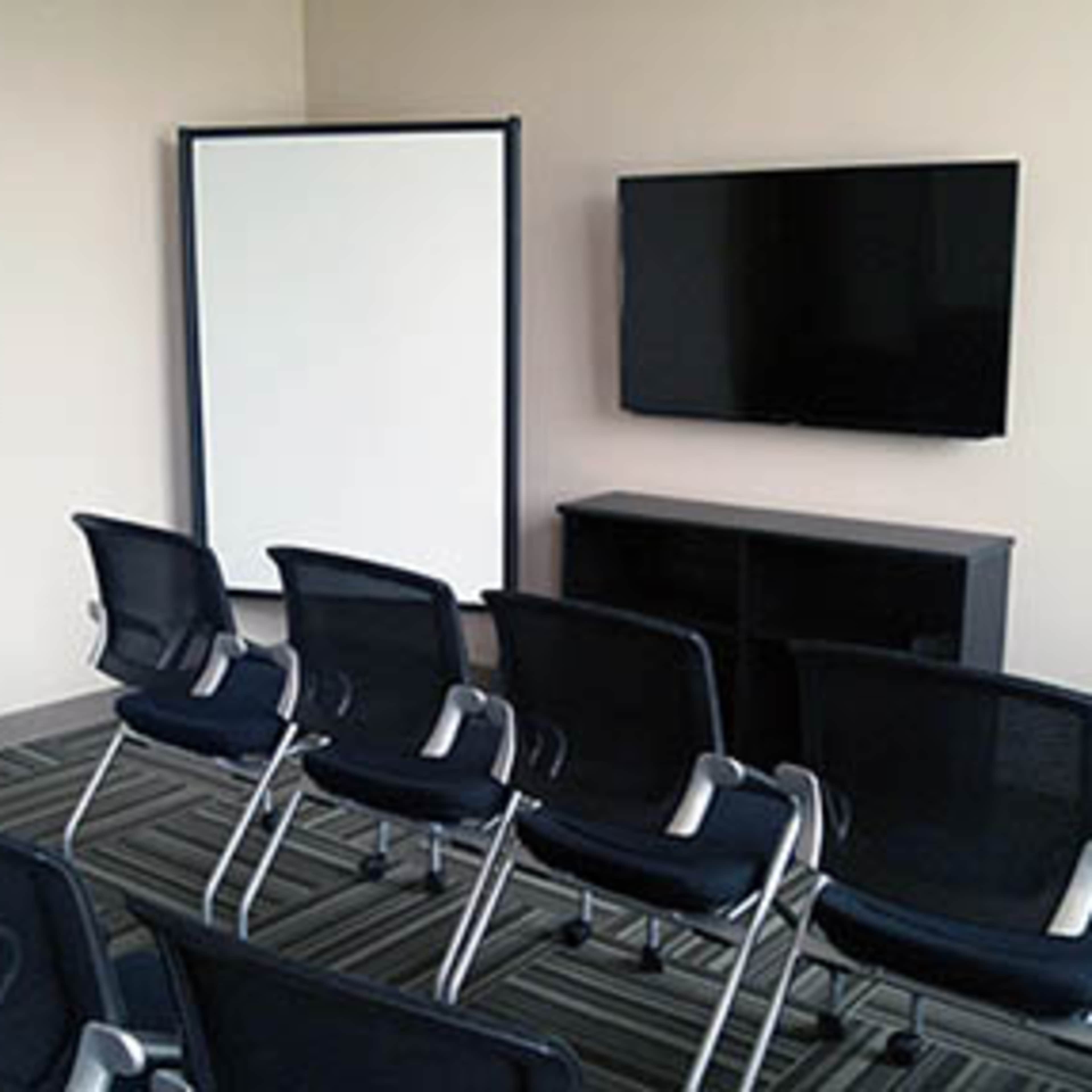 A meeting room is set up with several black chairs facing a wall-mounted television and a movable whiteboard.